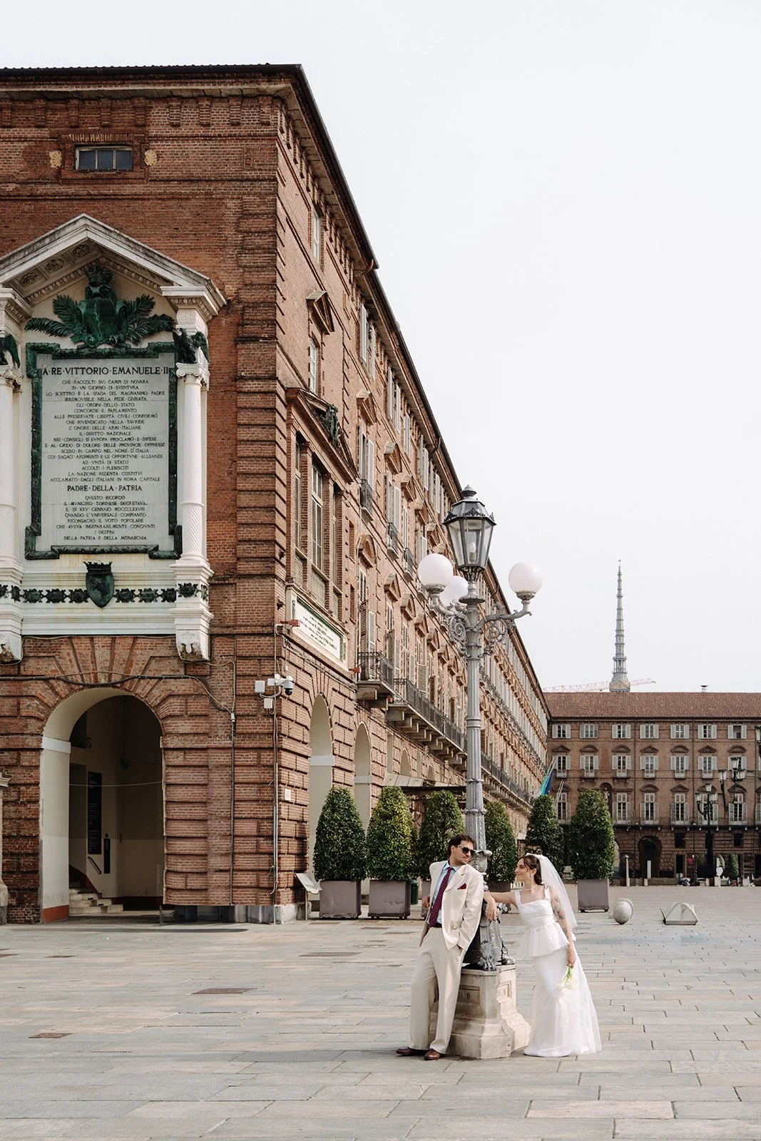 A bride and groom in wedding attire standing in an open plaza with historic brick buildings, street lamps, and potted plants in the background.