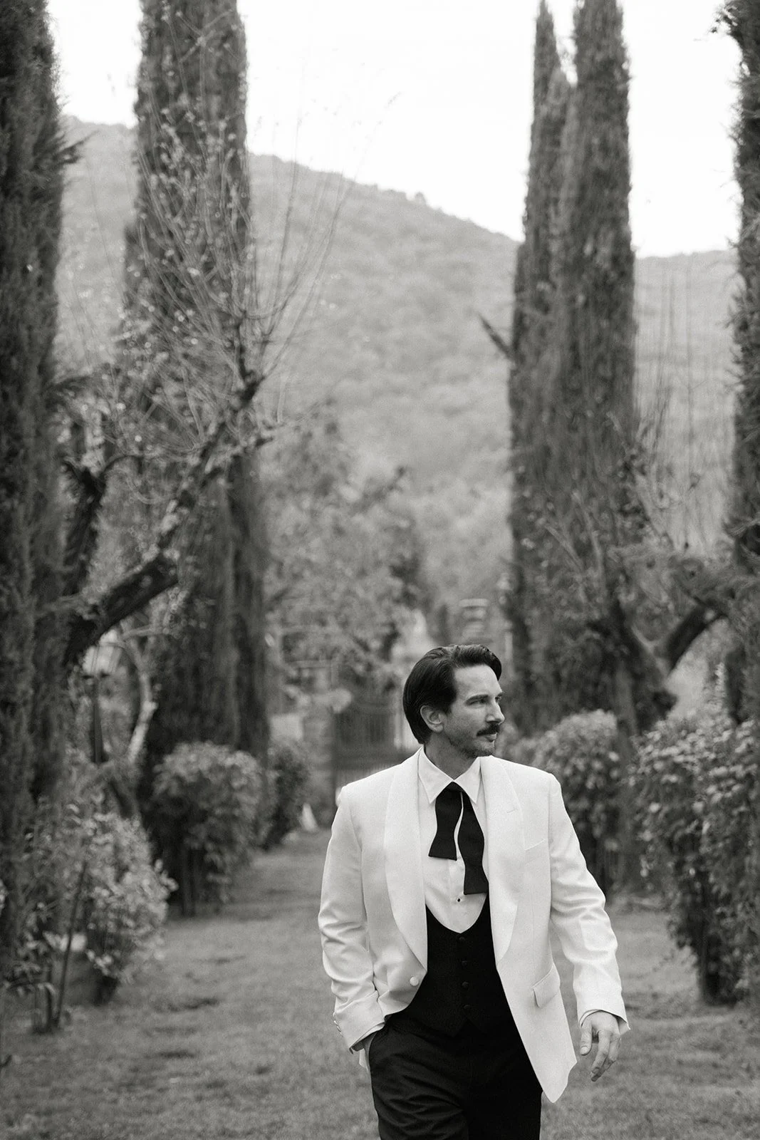 A man in a tuxedo jacket walking along a garden path lined with tall, leafless trees and bushes.