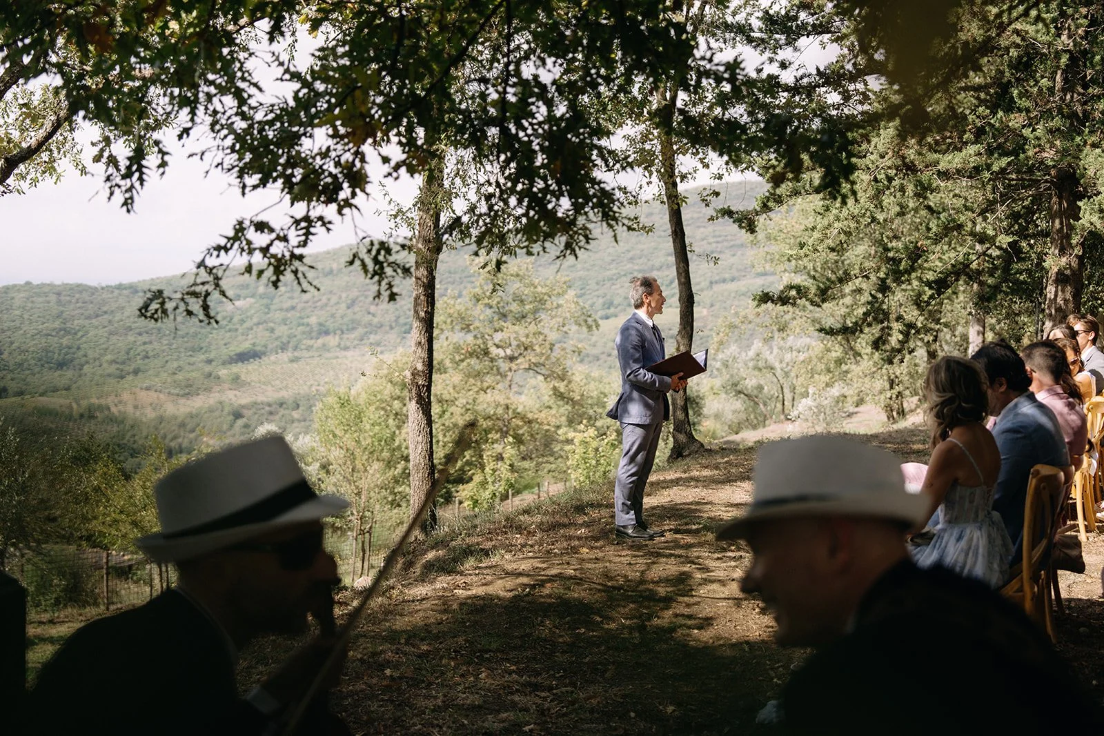 Man in a suit giving a speech at an outdoor wedding ceremony under trees, with guests seated and listening.