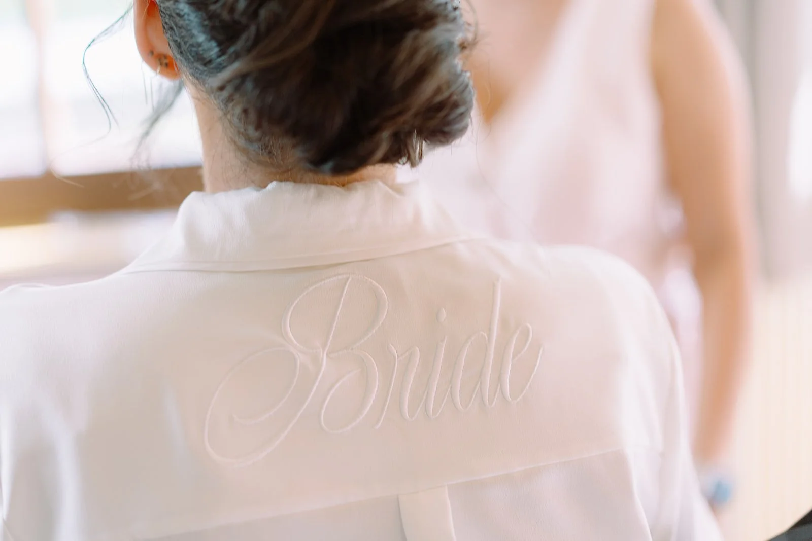 Back of a woman wearing a white robe with the embroidered name 'Bride' on the back. Her dark hair is styled up, and she is facing away from the camera.