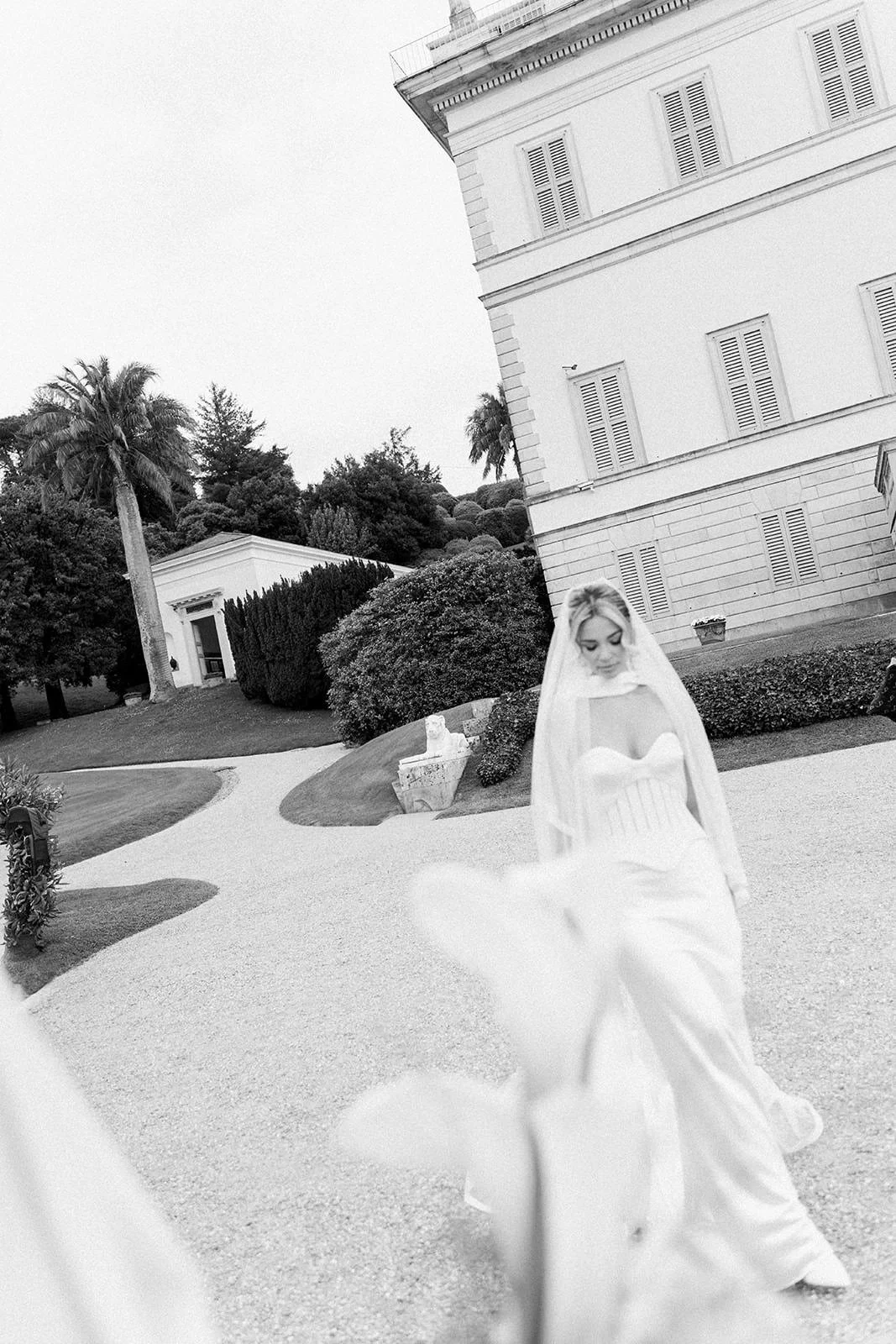 Black and white photo of a bride in a white wedding dress and veil walking outside in front of a large building with shutters, surrounded by trees and shrubs.