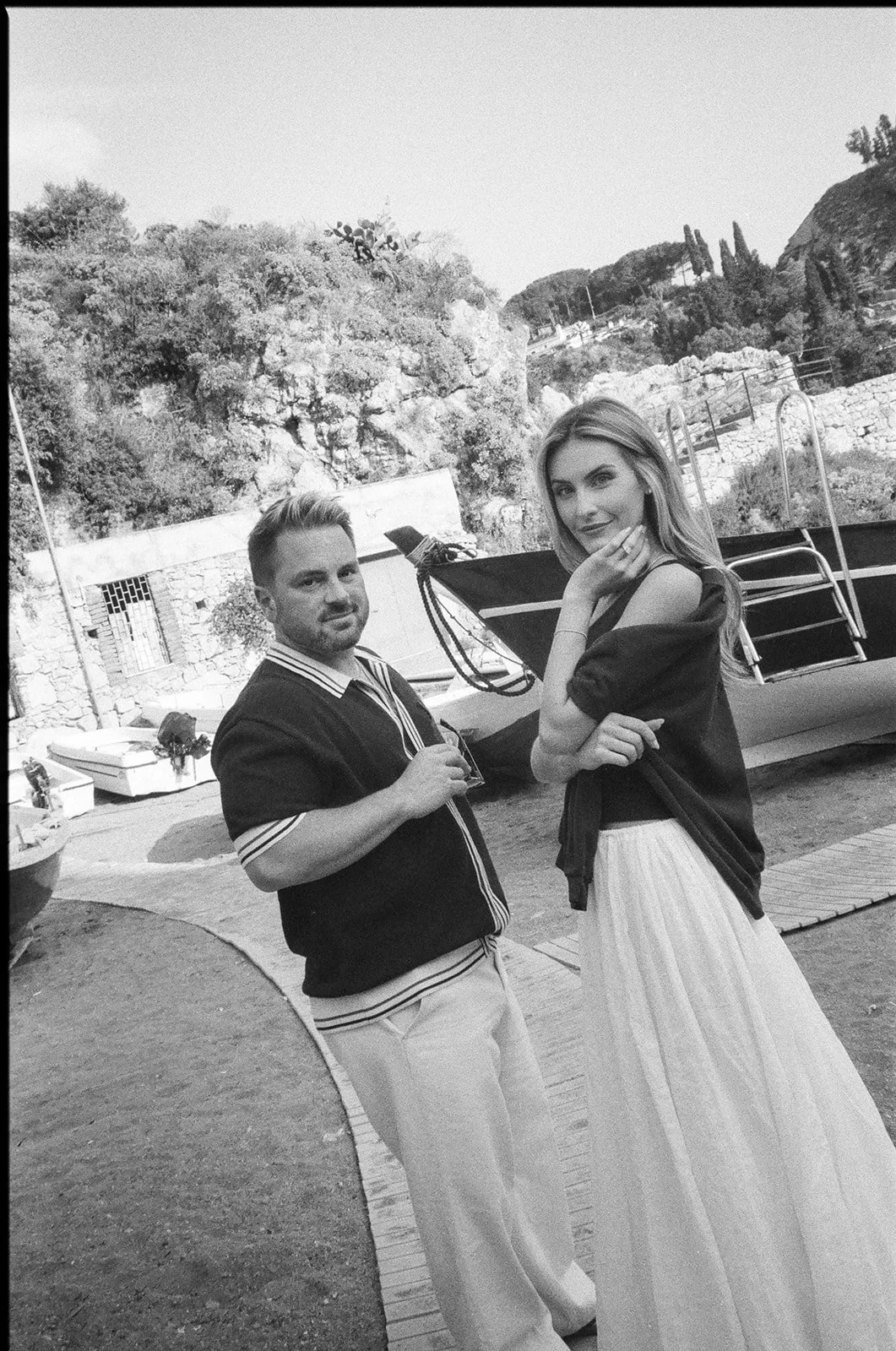 Two people, a man and a woman, standing outdoors near a boat with rocks and trees in the background.