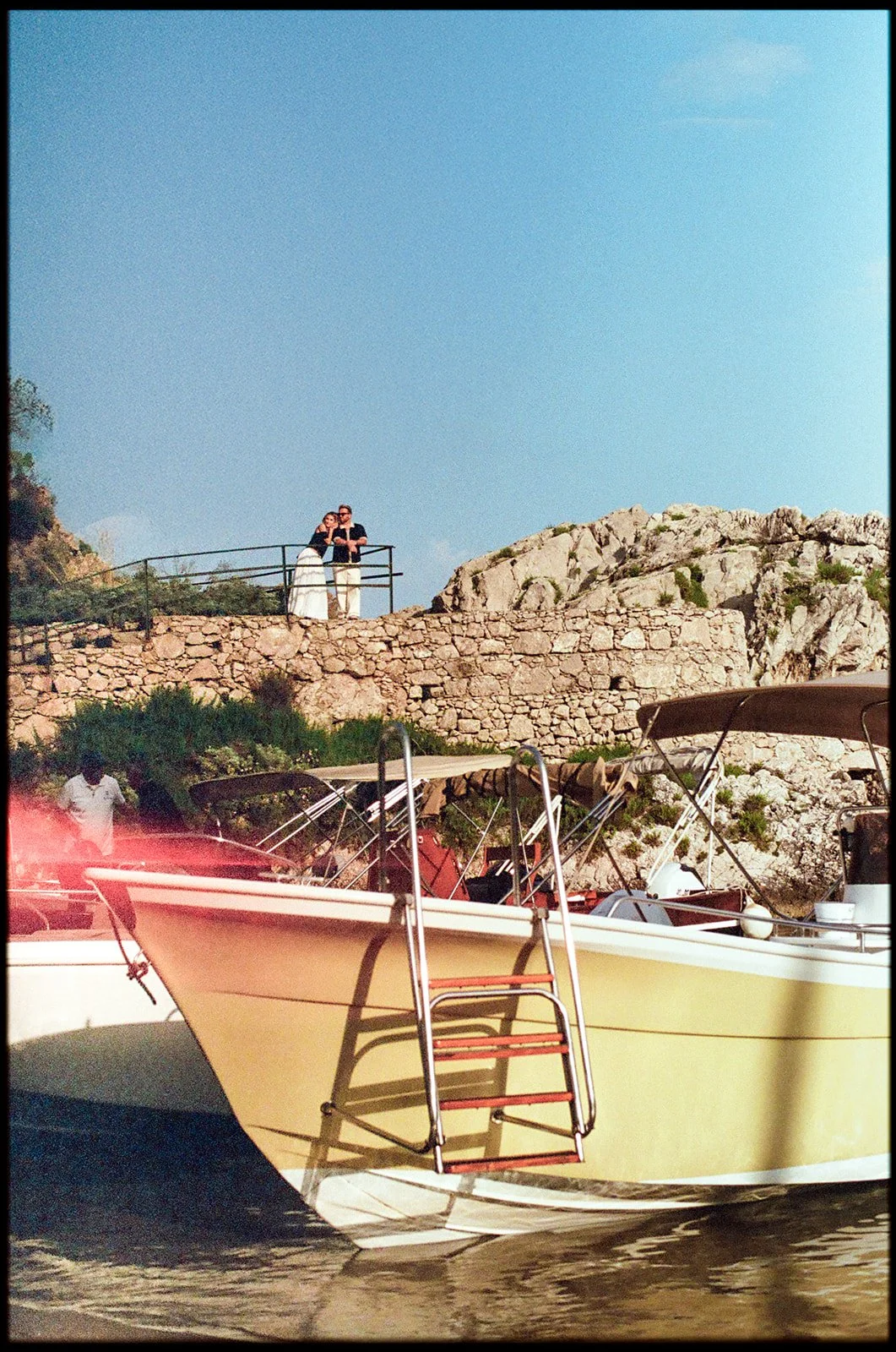 A boat docked on the water with a stone wall and rocky hillside in the background. Two people are standing on a viewing platform above, looking out over the scene.
