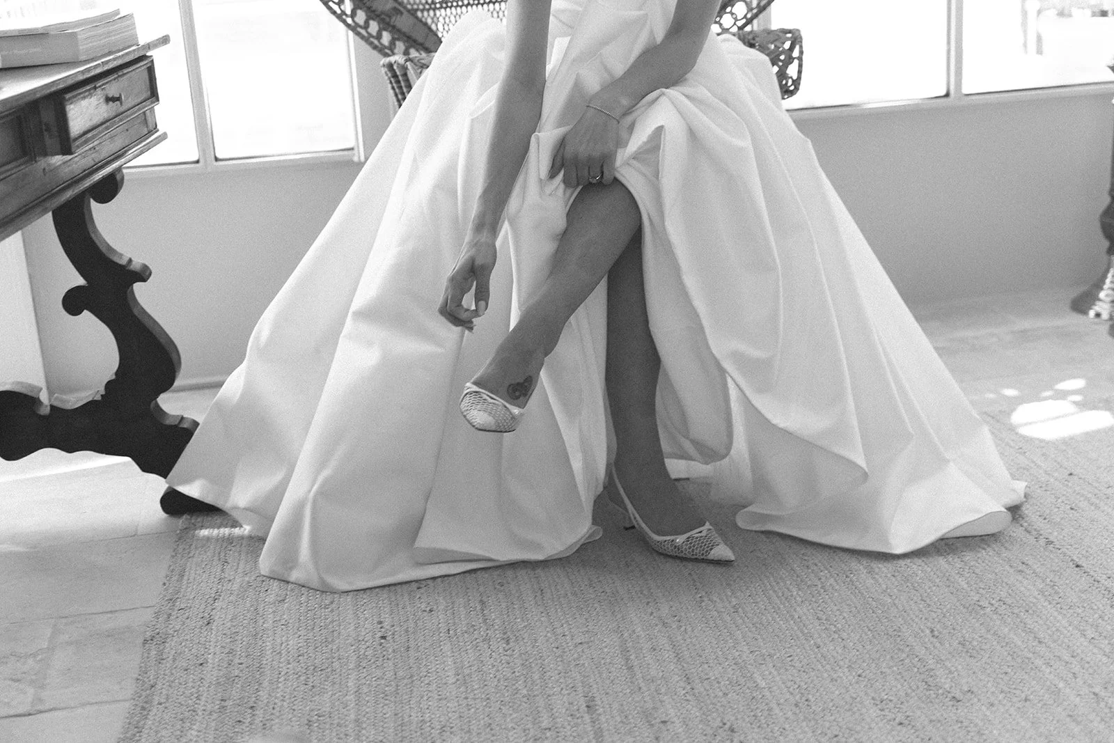 A person, likely a bride, is wearing a wedding dress and pointed high heels. She is tentatively lifting her dress to show her leg, revealing a tattoo and her shoe. The setting appears to be indoors near a window with sunlight coming through.