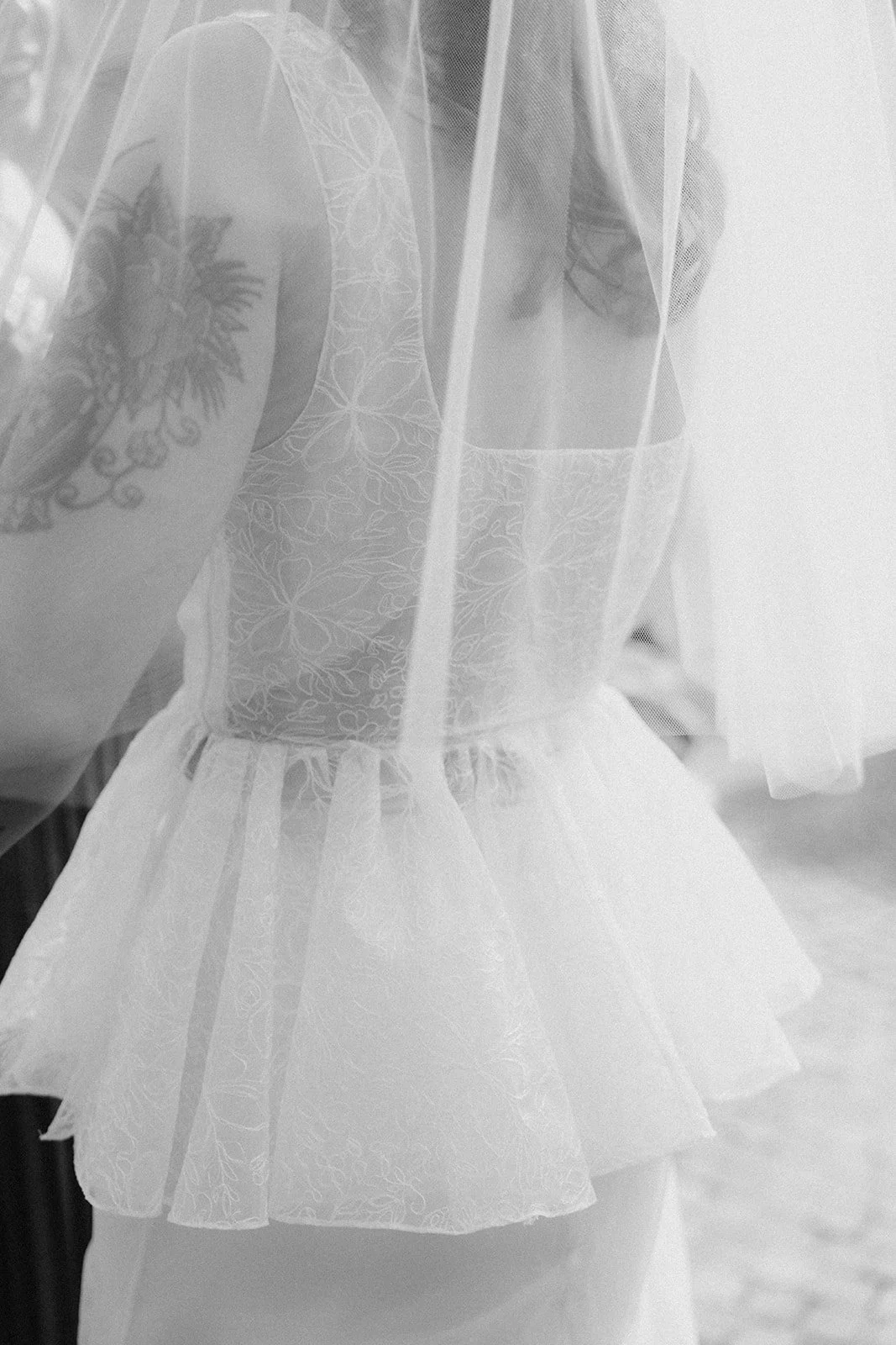 A woman wearing a lace off-shoulder dress and veil, with a tattoo on her arm, standing behind a sheer curtain.