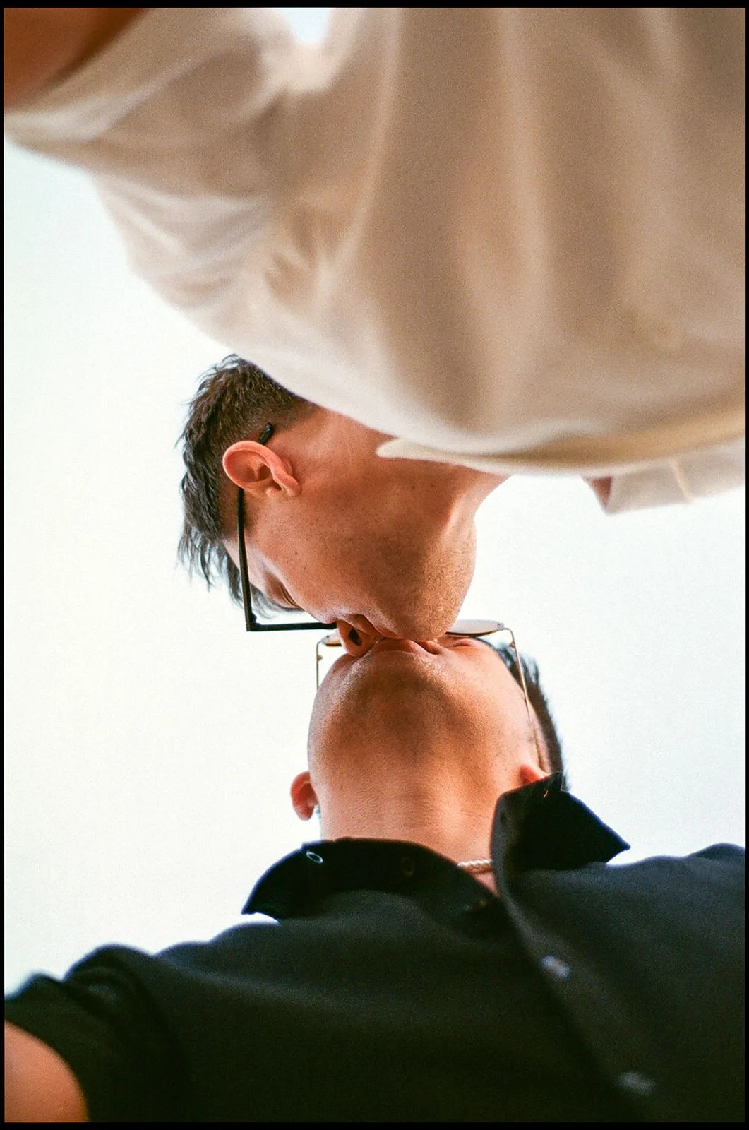 A man in a beige shirt kissing a woman with short hair, both wearing glasses, taken from below with a white background.