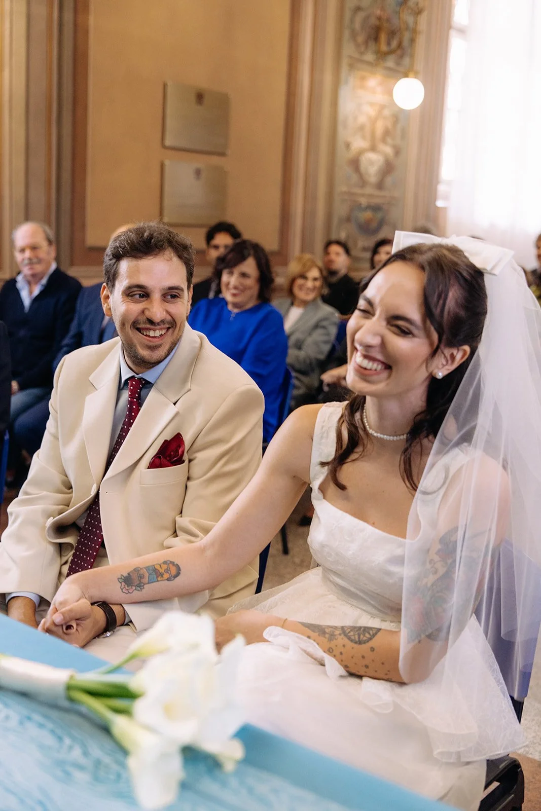 A bride and groom sitting hand in hand during their wedding ceremony, smiling at each other, with guests in the background in a decorated church interior.
