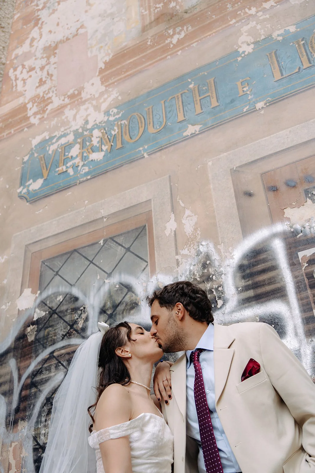 A bride and groom kissing in front of a painted wall with the words 'Vermont South'. The bride is wearing a white off-the-shoulder wedding dress and a veil, and the groom is wearing a beige suit with a red pocket square and a polka dot tie.