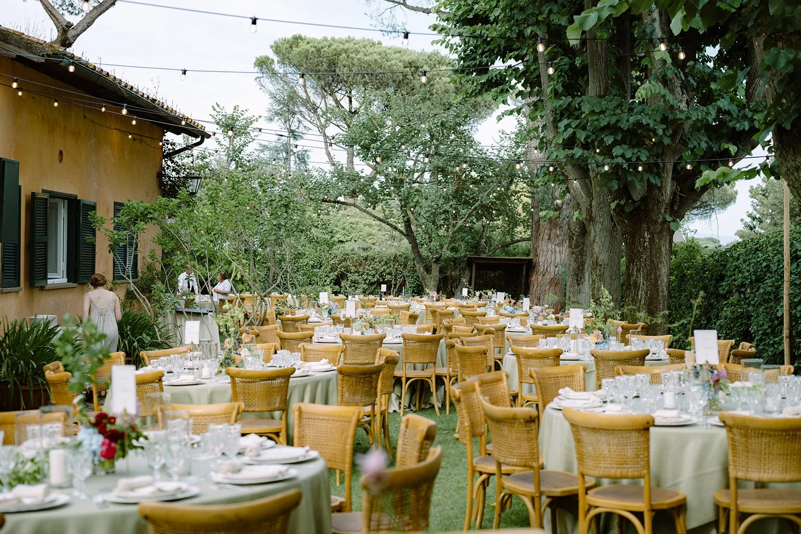 Outdoor wedding reception setup with tables and chairs under string lights, surrounded by trees and greenery.