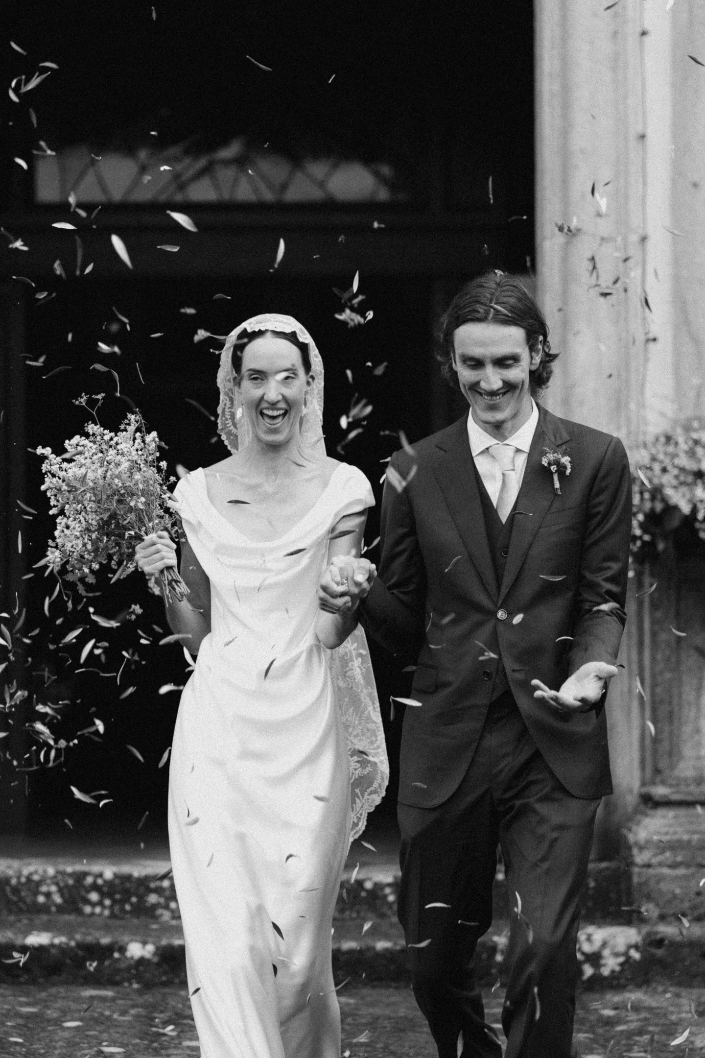 A bride and groom smiling joyfully as they walk through falling confetti outside, celebrating their wedding.