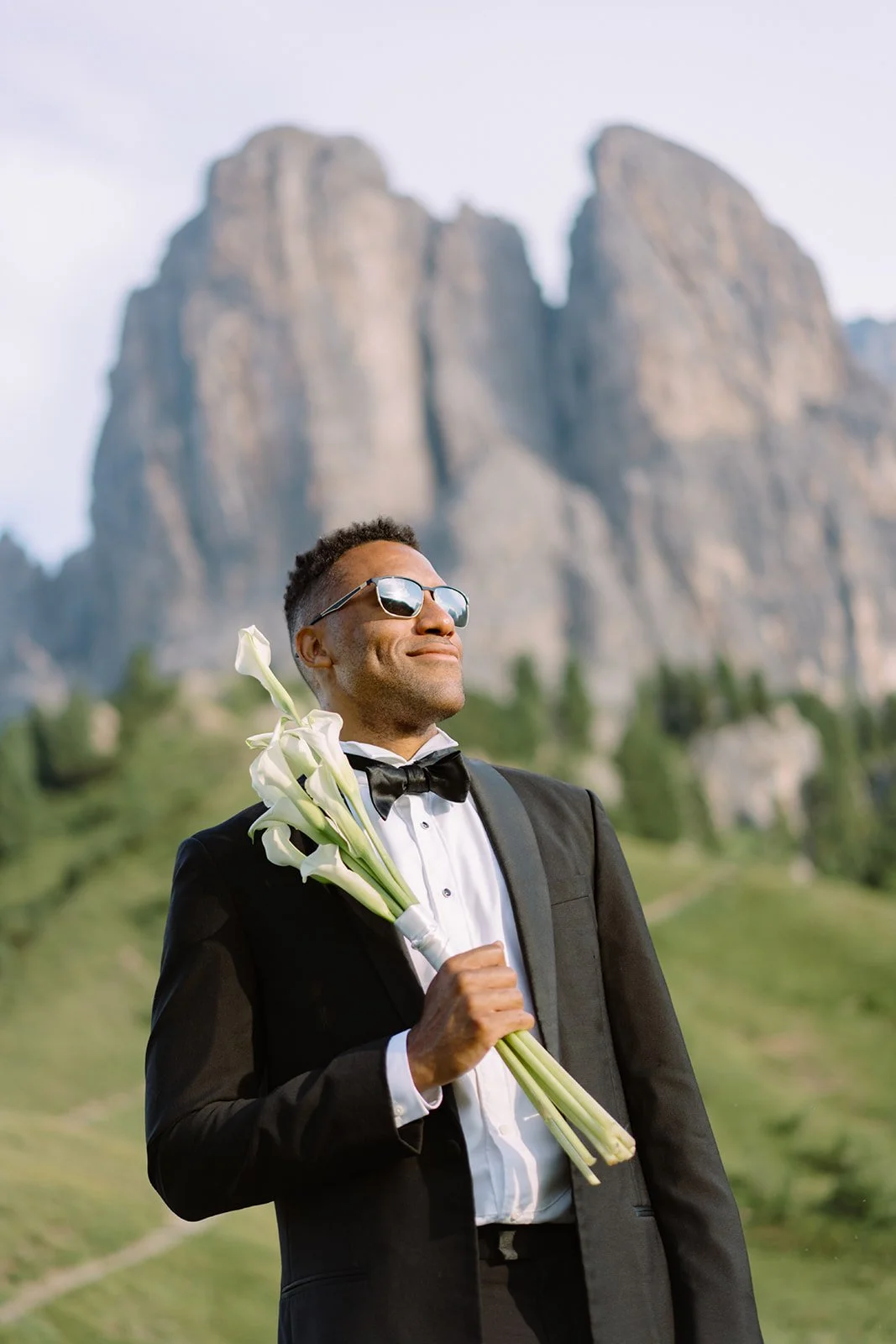 A man in a tuxedo and sunglasses holding a bouquet of white calla lilies outdoors with mountains and greenery in the background.