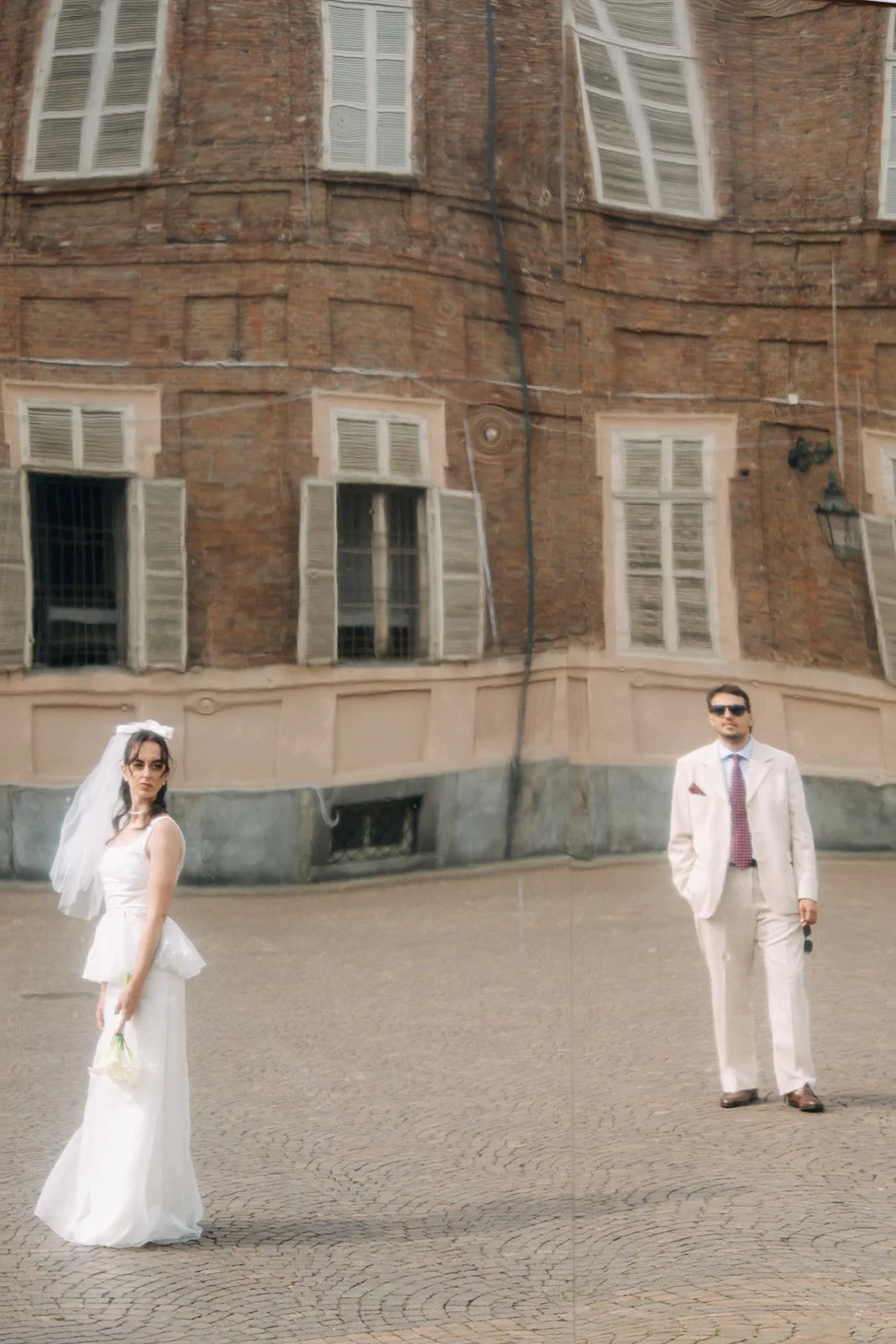 A bride in a white wedding dress holding a small bouquet of flowers and a man in a light-colored suit with sunglasses standing in front of a building with reflective windows.
