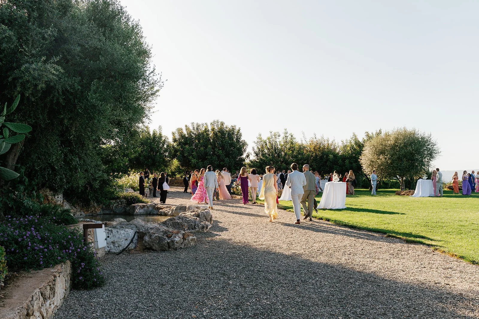 People attending an outdoor wedding reception in a garden, with guests walking on a gravel path surrounded by greenery.