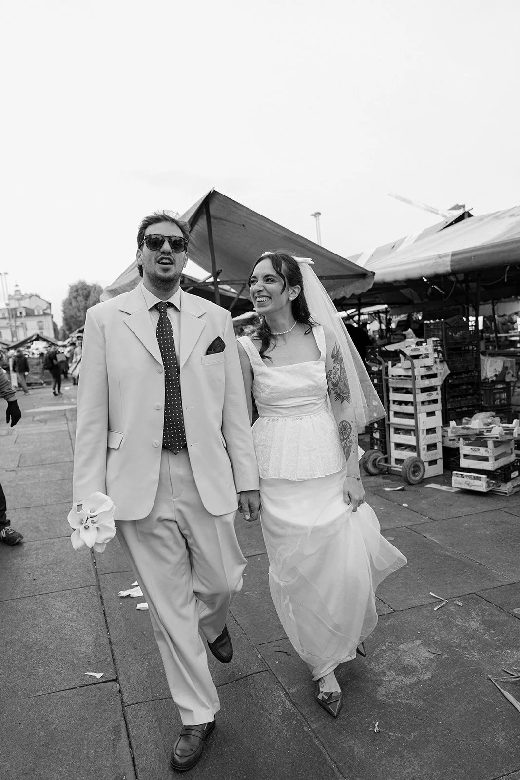 A man and woman in wedding attire walking outdoors, smiling and laughing. The man is wearing a light suit with sunglasses and a polka dot tie, holding flowers. The woman is dressed in a white wedding dress with a veil, tattoos visible on her arms, an