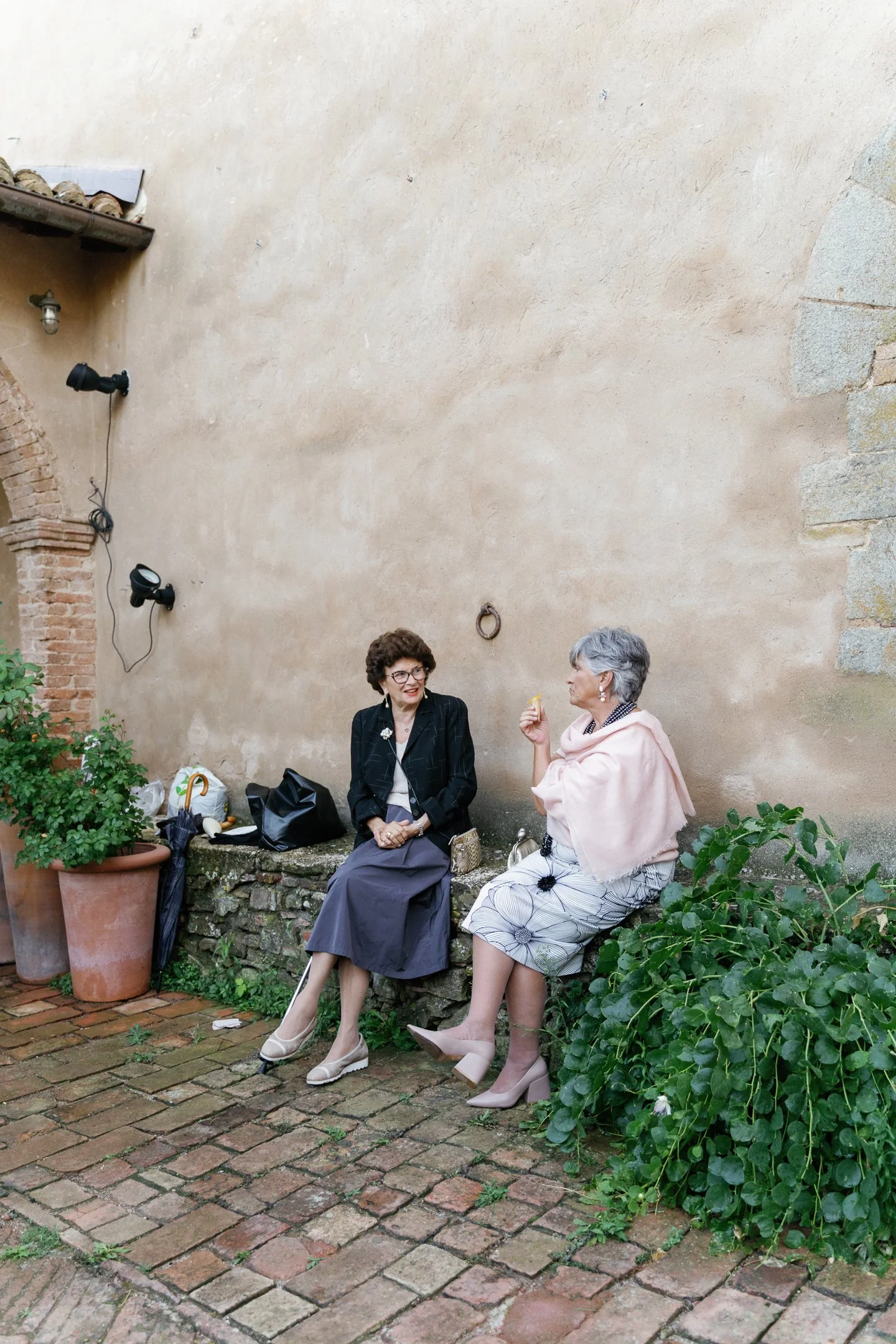 Two women sitting on a stone ledge and talking in an outdoor space with brick and stucco walls, potted plants, and old brick pavement.