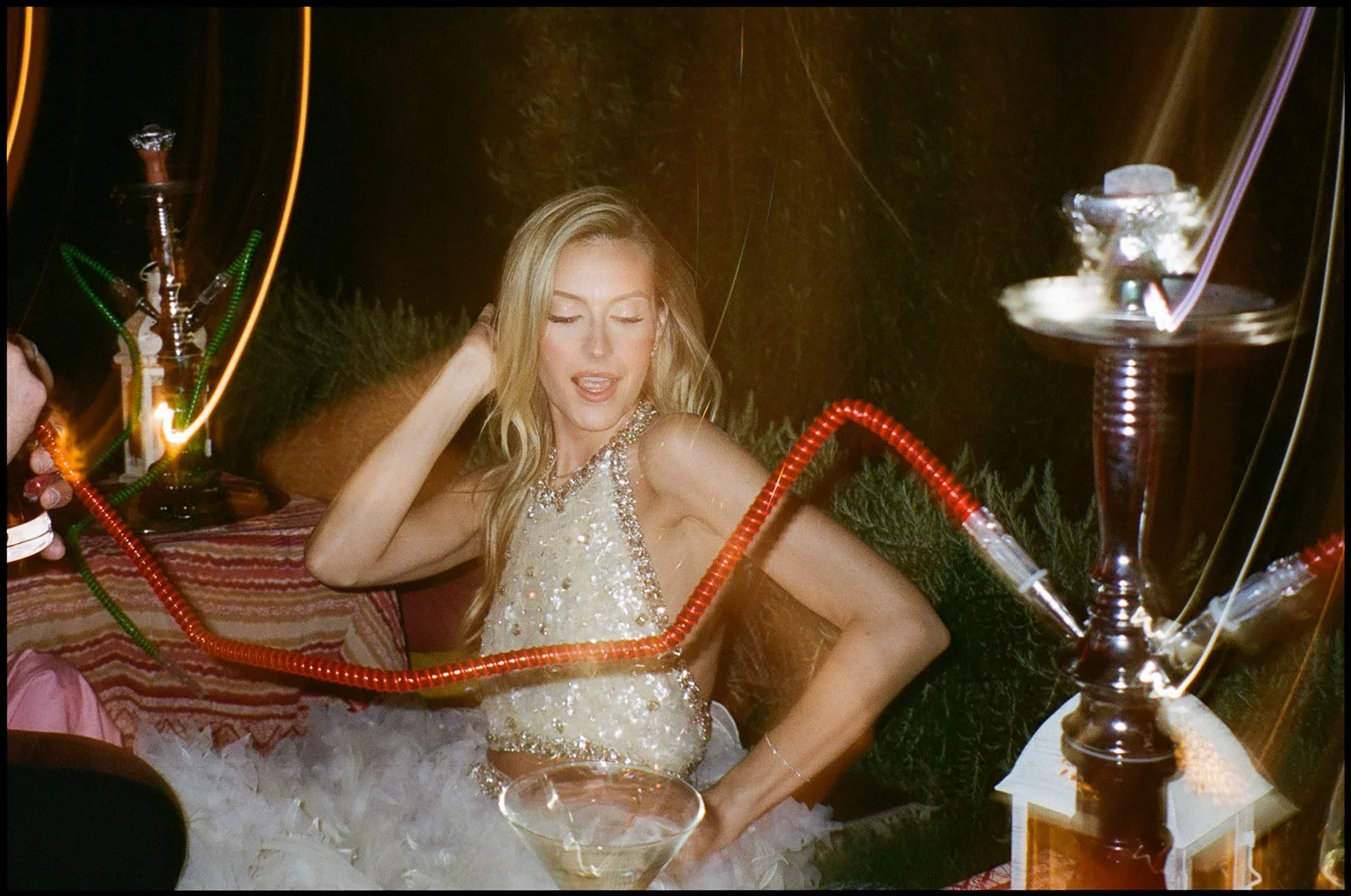 A woman in a sparkly dress sitting on a cushioned surface, surrounded by hookah pipes and a cocktail glass, with a relaxed expression at a party.