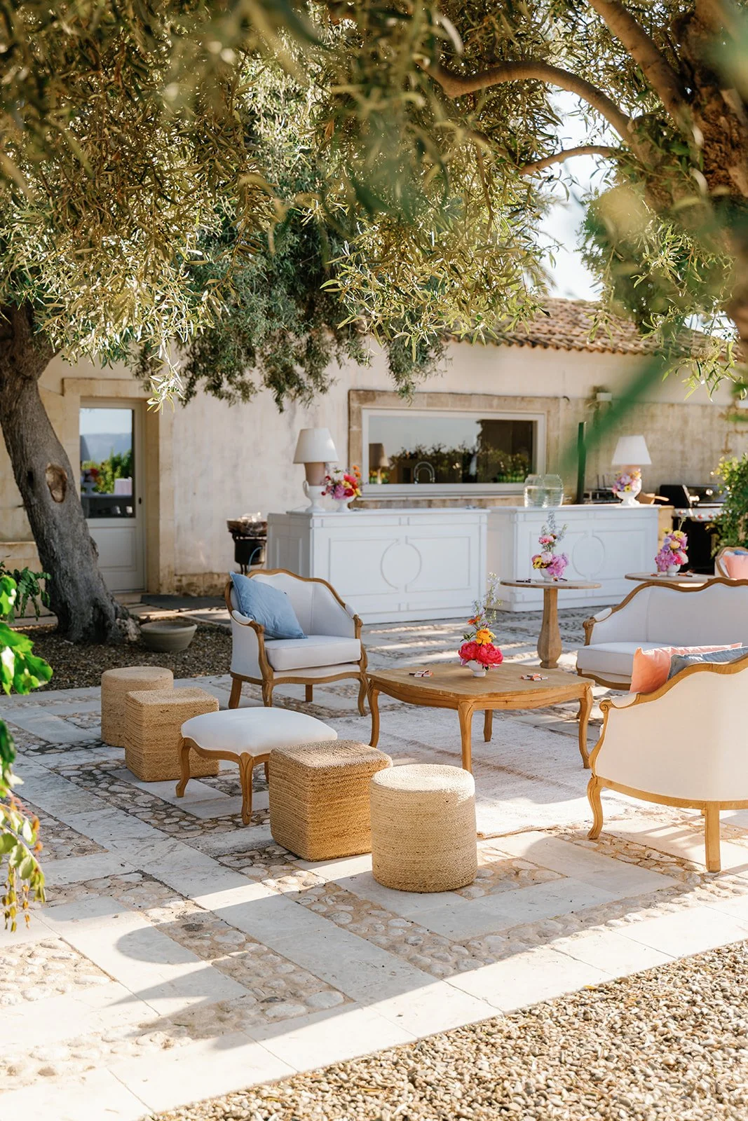 Outdoor patio with white and wooden furniture, colorful flowers in vases, trees, and a building in the background.