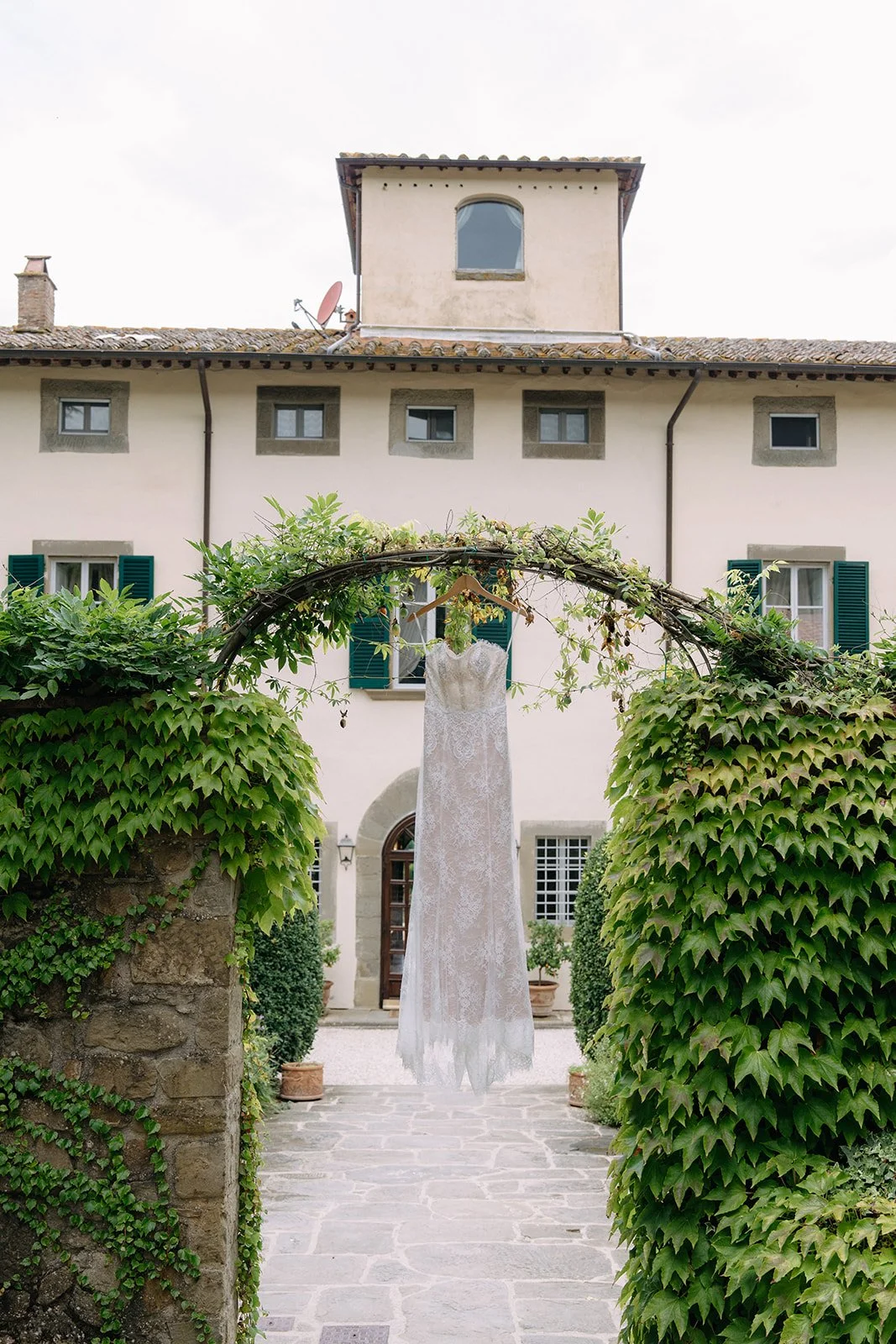 A white wedding dress hanging on a hanger from a vine-covered archway in front of a European-style building with green shutters.