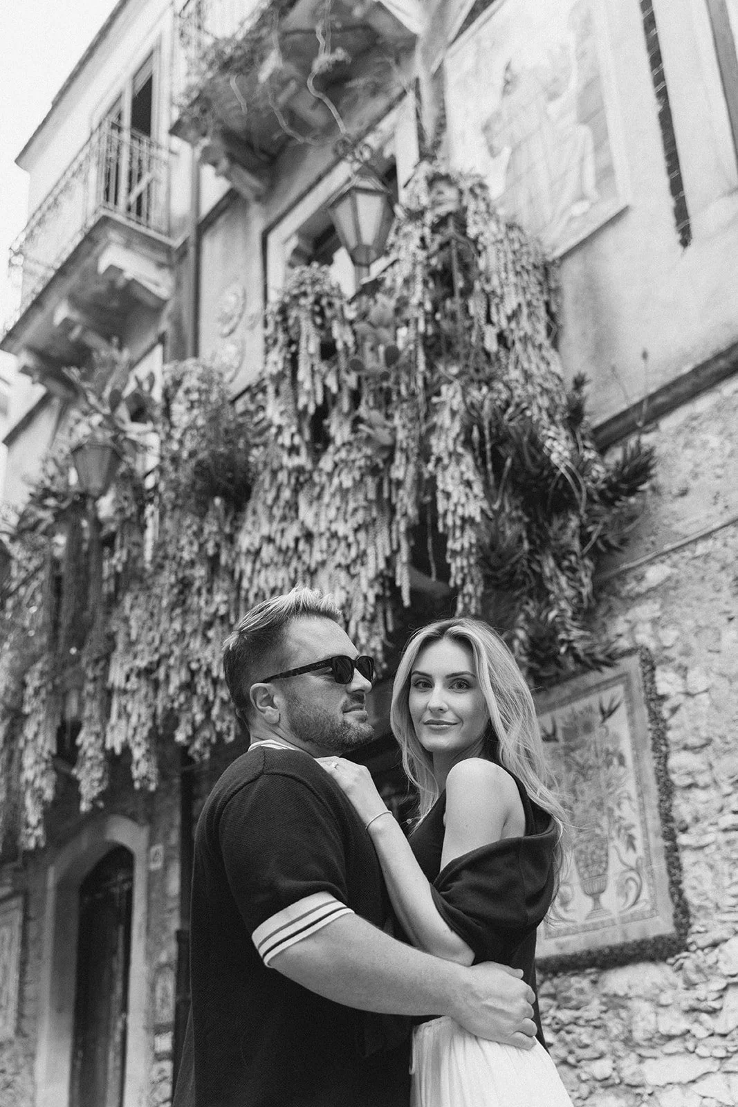 A couple posing for a photo in front of a decorative building with ivy and hanging ornaments, in black and white.