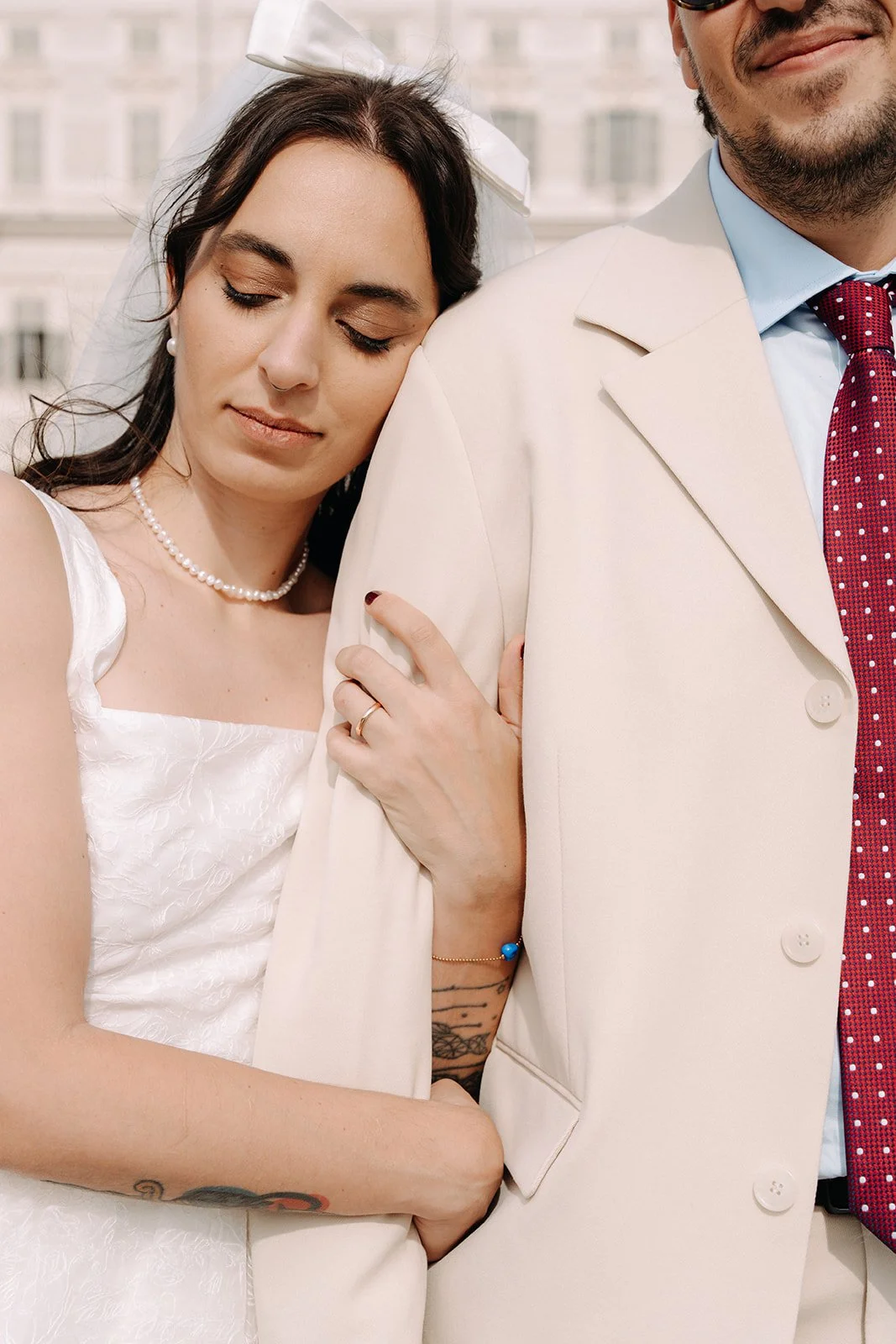 A woman in a white dress with a pearl necklace, with her eyes closed, leaning on a man's shoulder who is wearing a cream-colored suit and a red polka dot tie.