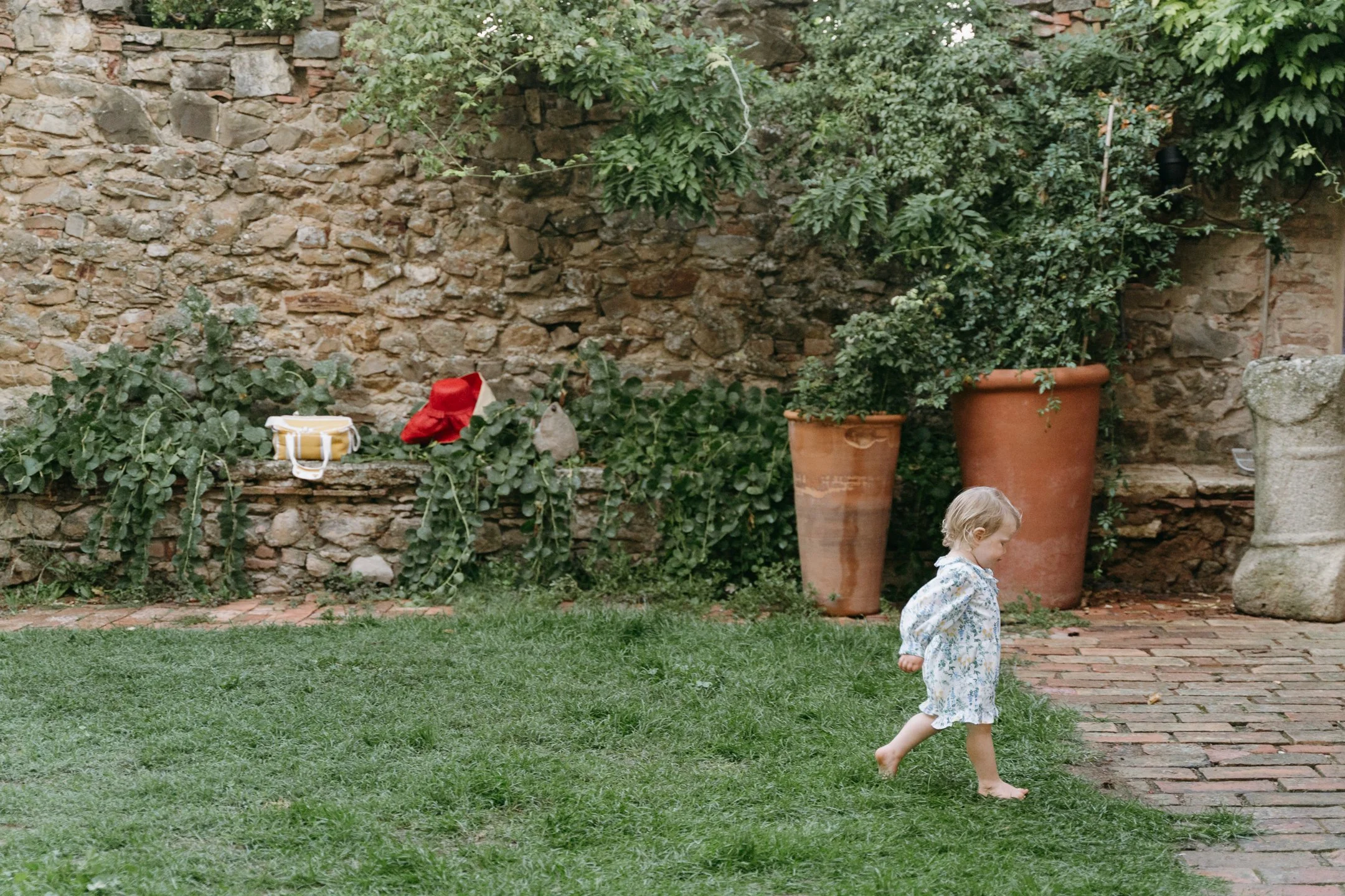 A toddler walking barefoot on grass and brick paving in a garden, with large terracotta pots and a stone wall in the background.