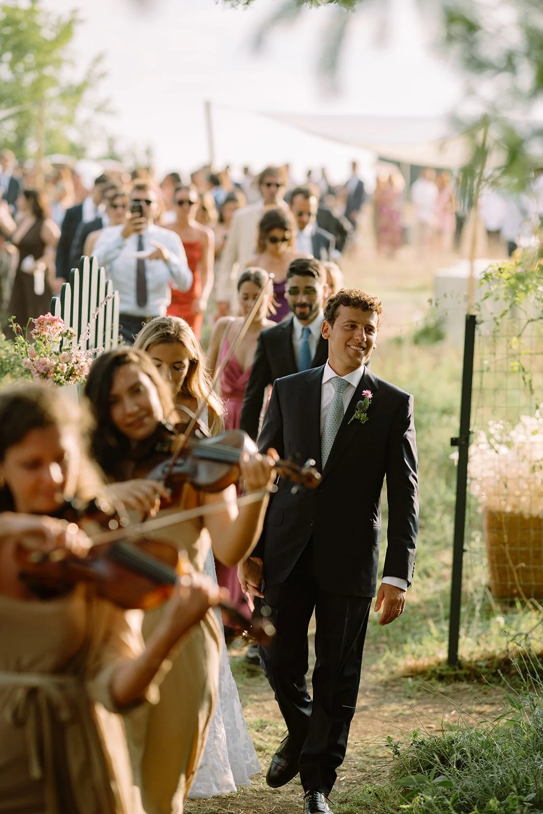 A smiling groom in a black suit walking past musicians playing violins at an outdoor wedding ceremony