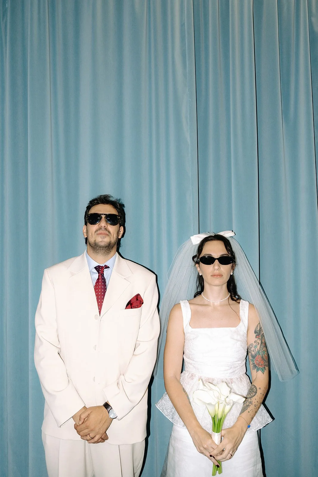 A man and a woman in wedding attire standing side by side against a blue curtain. Both are wearing sunglasses, and the woman is holding a bouquet of white calla lilies.