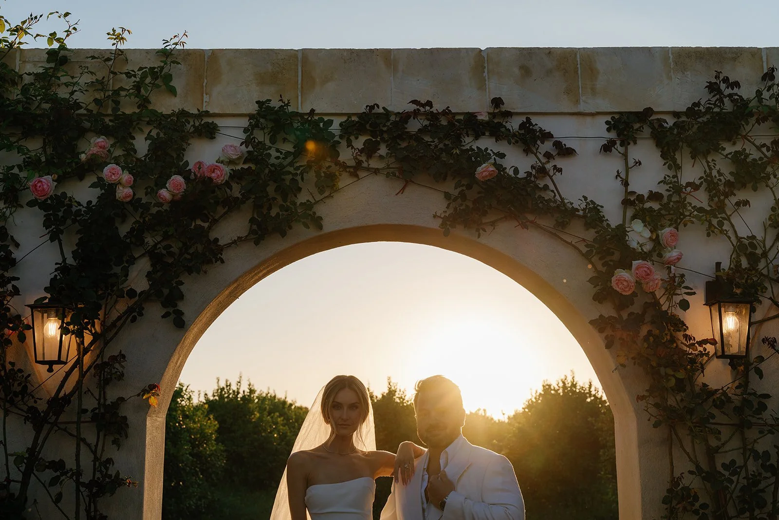 A bride and groom standing under an archway decorated with pink roses and climbing vines, with the setting sun behind them.