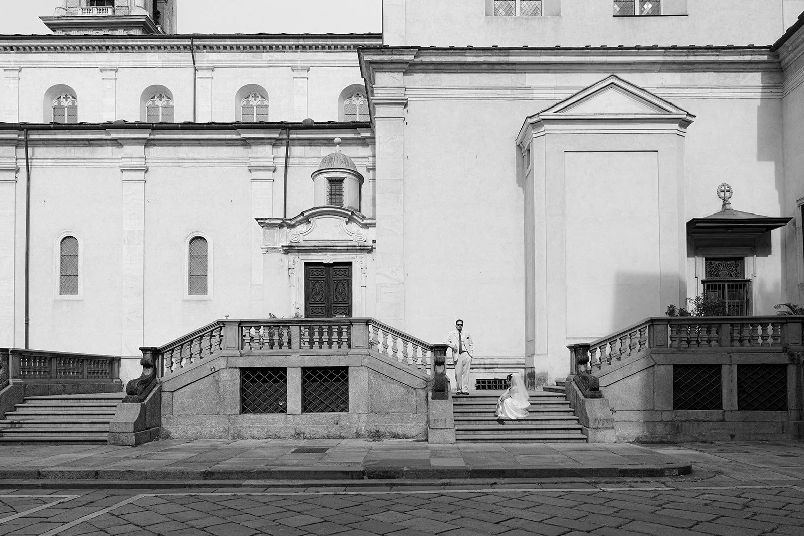 A black and white photograph of a grand building with stairs leading up to an entrance. Two people are on the stairs: a man in a suit standing and a woman in a wedding dress sitting on the stairs. The building features arched windows, a small turret,