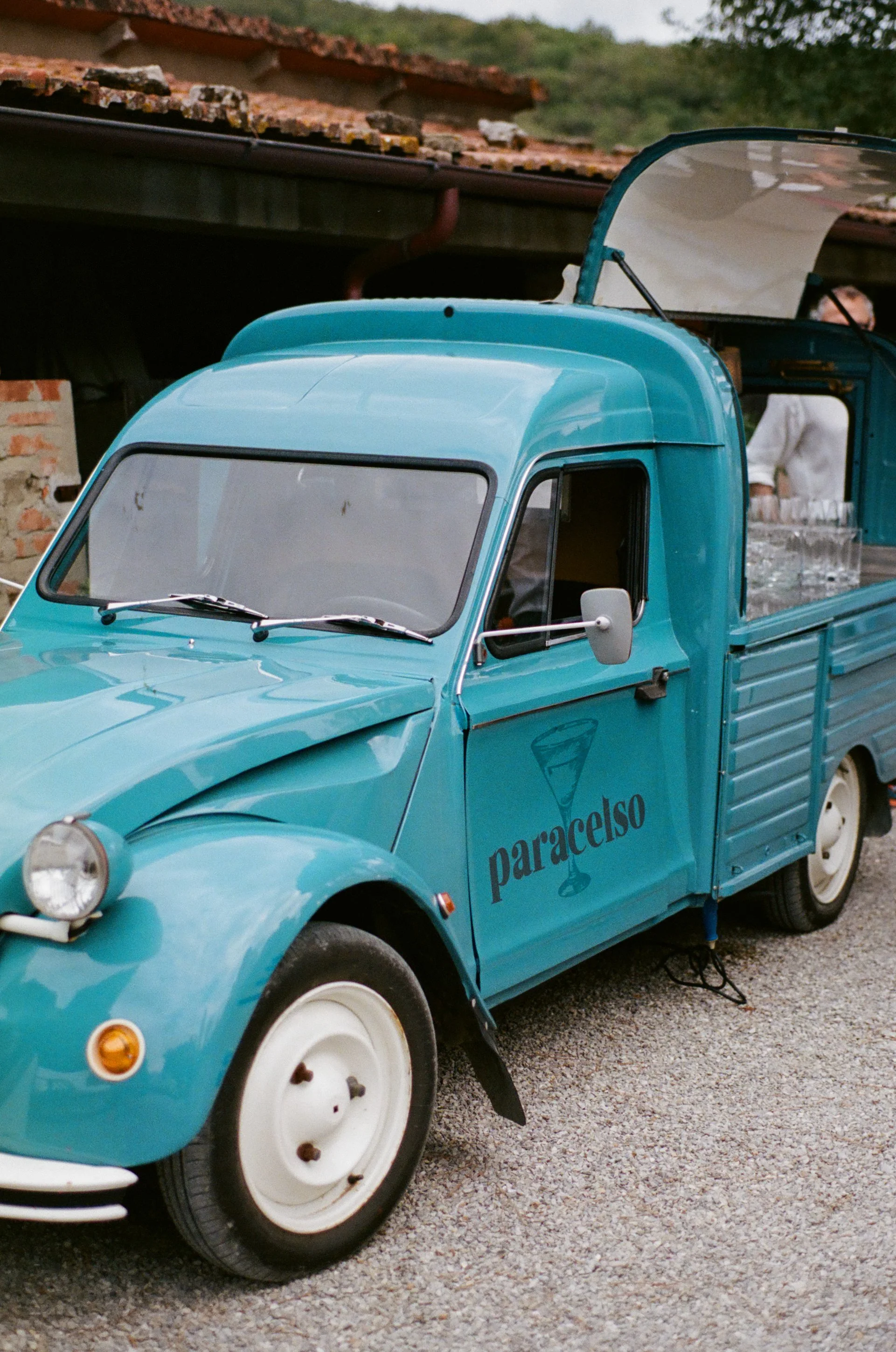 A vintage blue Piaggio Ape three-wheeled vehicle with the words "paracelso" and a cocktail glass logo on the door, parked on gravel.