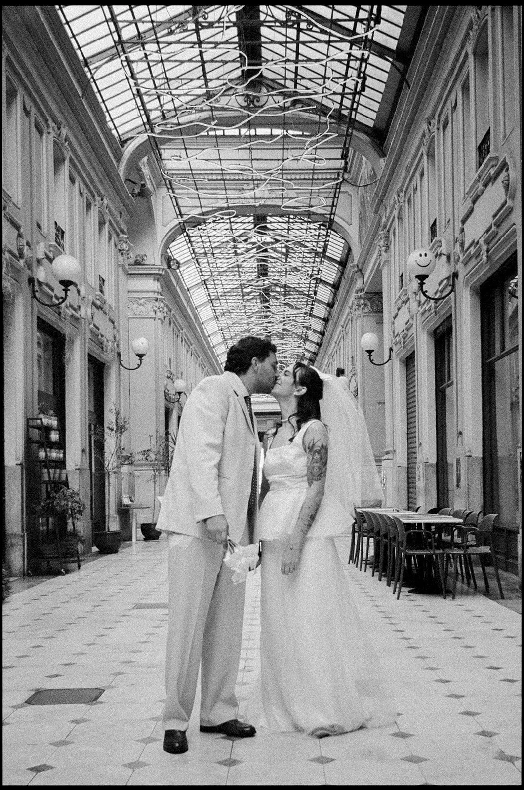 A black and white photo of a bride and groom sharing a kiss in a vintage indoor arcade with glass ceilings and ornate wall details.