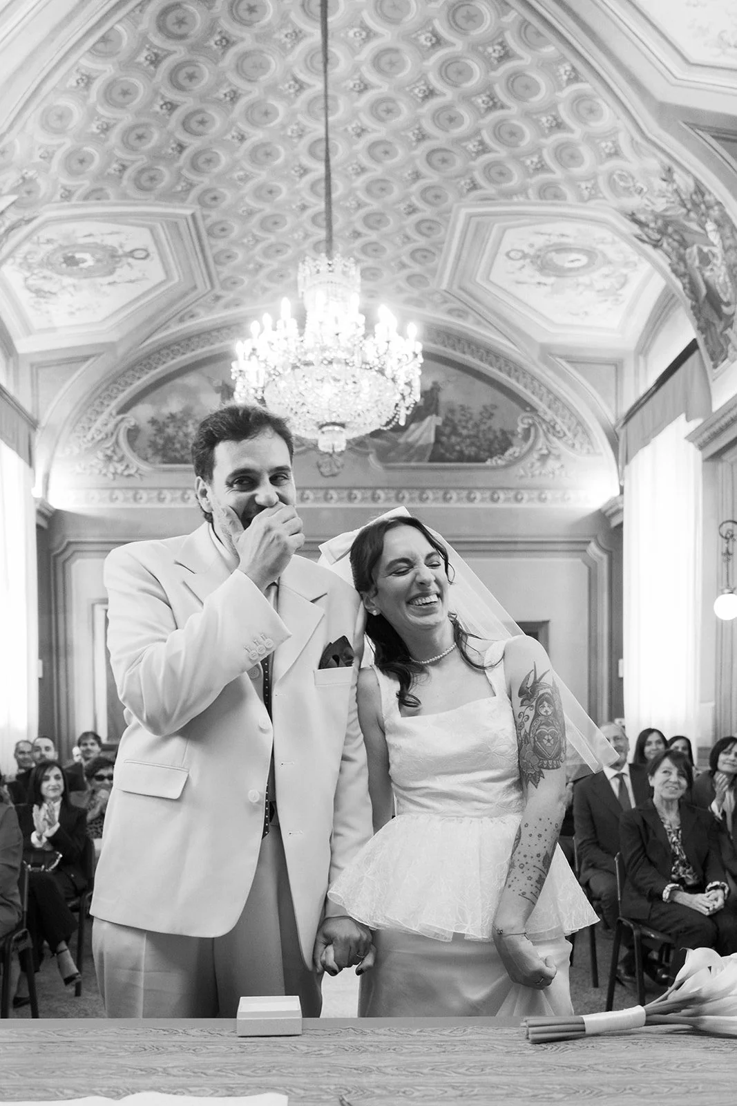 A black and white photo of a bride and groom standing hand in hand during their wedding ceremony in a grand ornate hall with a chandelier. The bride is smiling and looking joyful, while the groom covers his mouth, possibly laughing or surprised. Gues