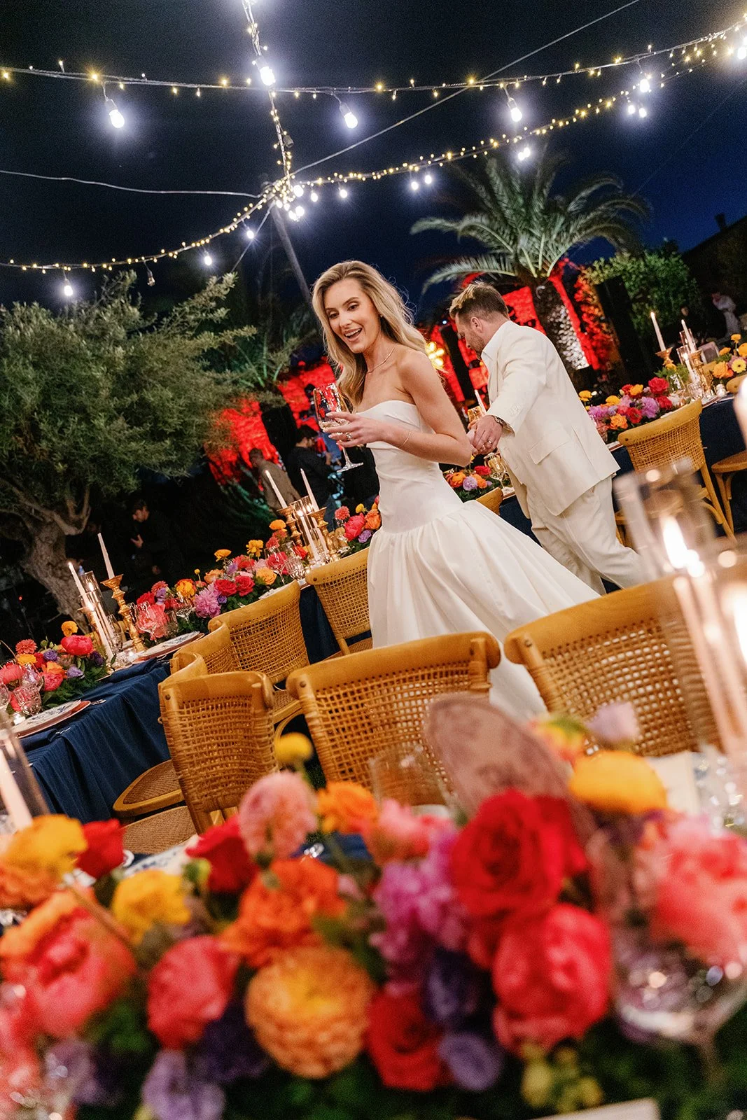 A bride in a white wedding dress dancing at a wedding reception under string lights at night, with tables decorated with colorful flowers in the background.