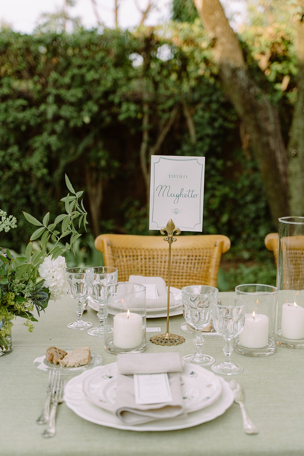 A beautifully set outdoor dining table with white plates, glassware, candles, and a floral centerpiece, with a sign that reads 'Tavolo Mughetto' amidst greenery and trees in the background.