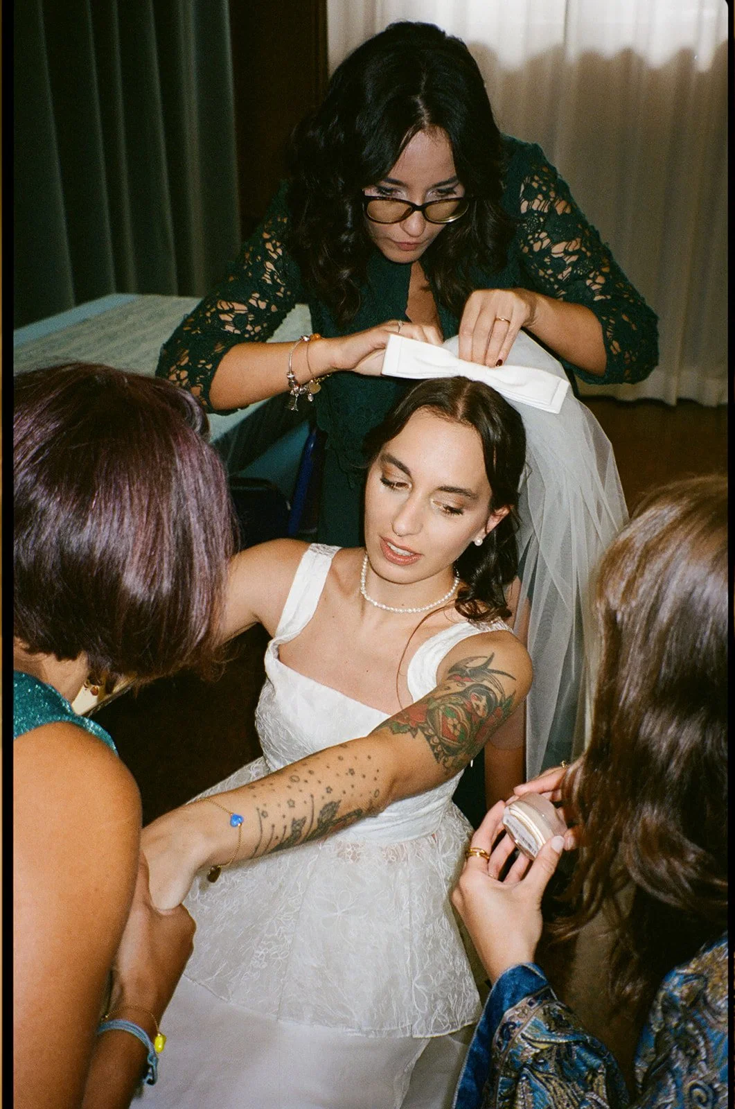 A bride with tattoos on her arm getting her wedding dress and makeup adjusted by friends and family in a room.