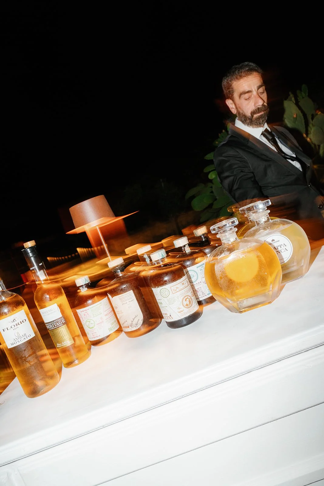 A man in a black suit with a beard and mustache is sitting at a table under dim lighting, with bottles of liquor, including some labeled 'Amarone' and 'Kappa Panna,' arranged on the table. The background is dark with foliage visible.