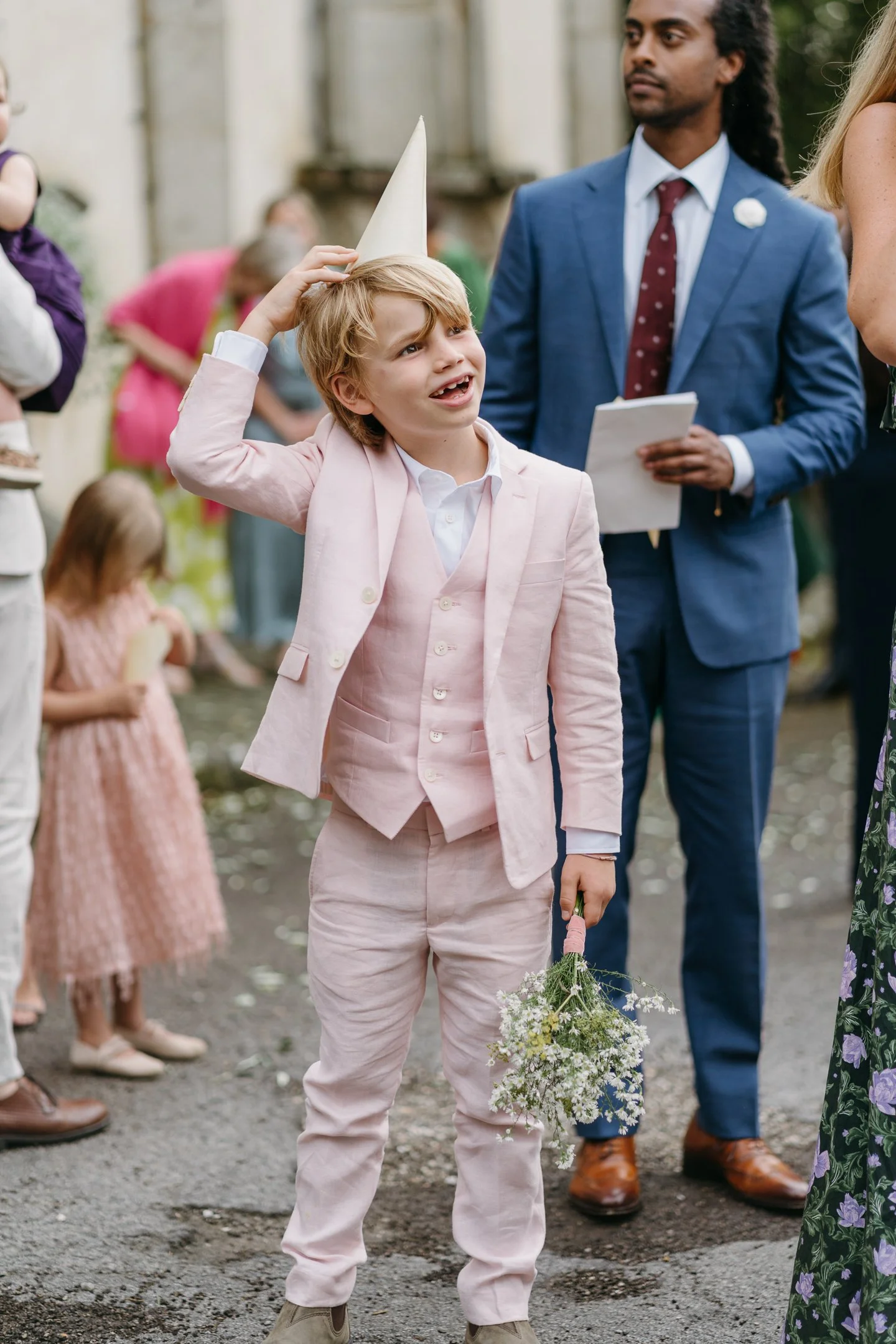 A young boy in a pink suit and vest, holding a bouquet of flowers, is gesturing up with his hand and appearing upset at an outdoor gathering. He wears a party hat, and people are standing around him, some holding children, with a man in a blue suit a