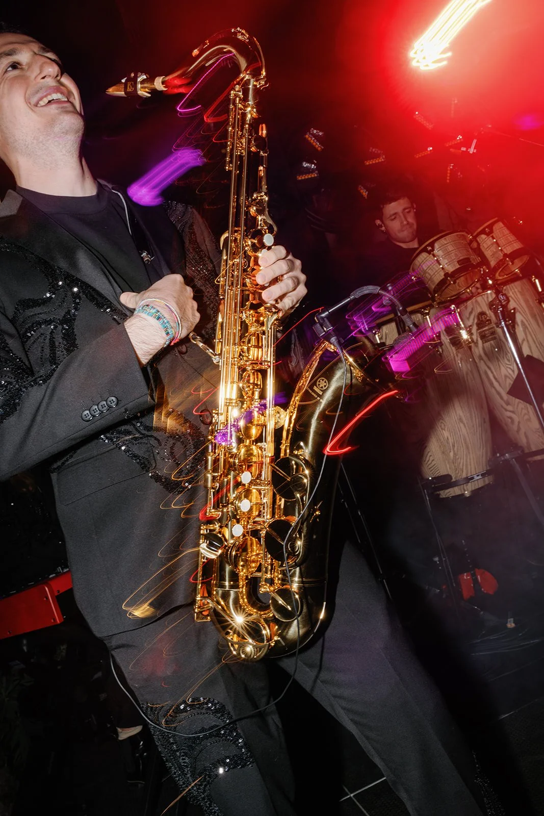 A musician playing a saxophone on stage with colorful lights and a percussionist in the background.