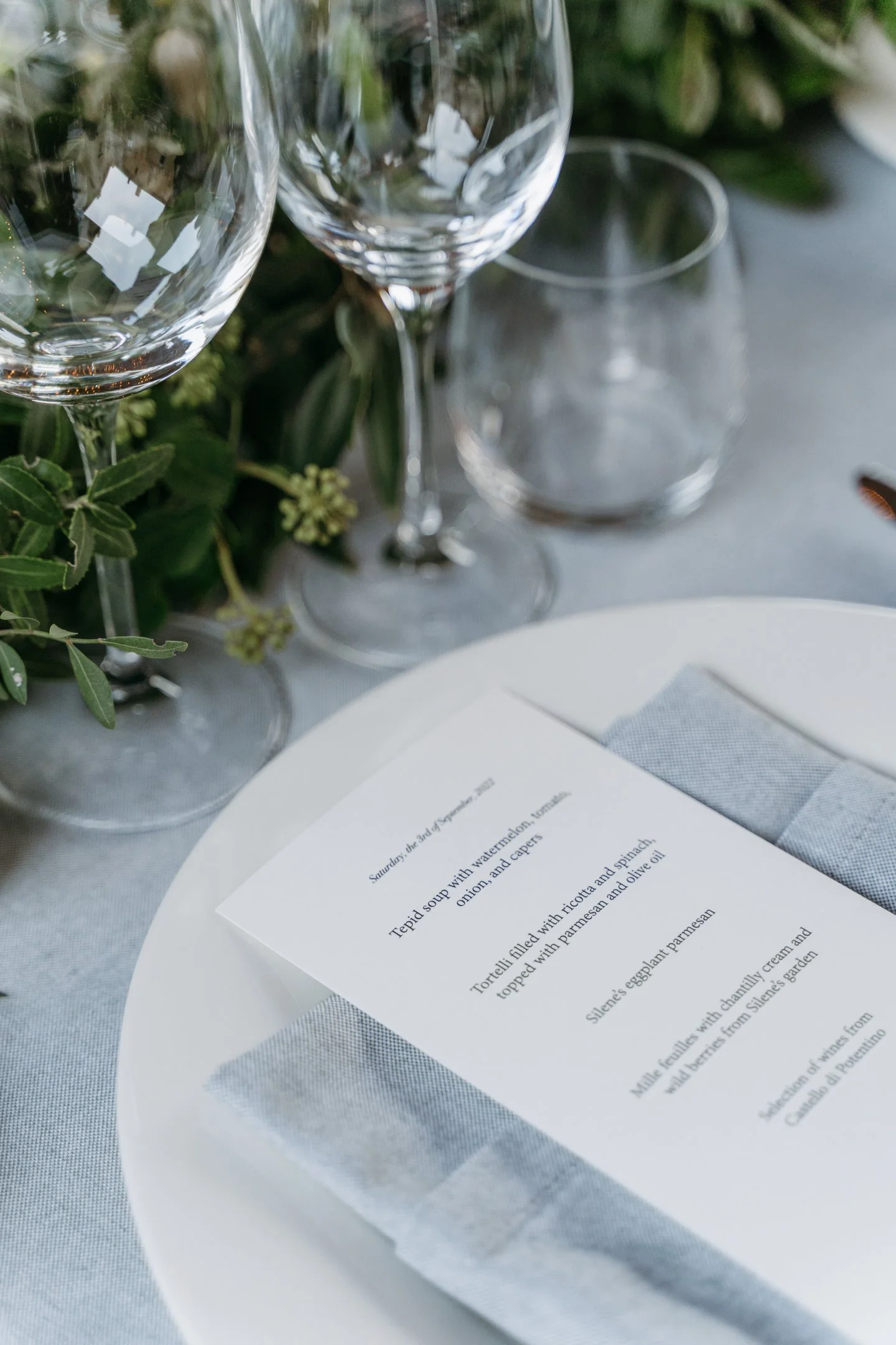Elegant table setting with clear wine glasses, a white plate, a gray cloth napkin, and a printed menu. Green foliage decorates the background.