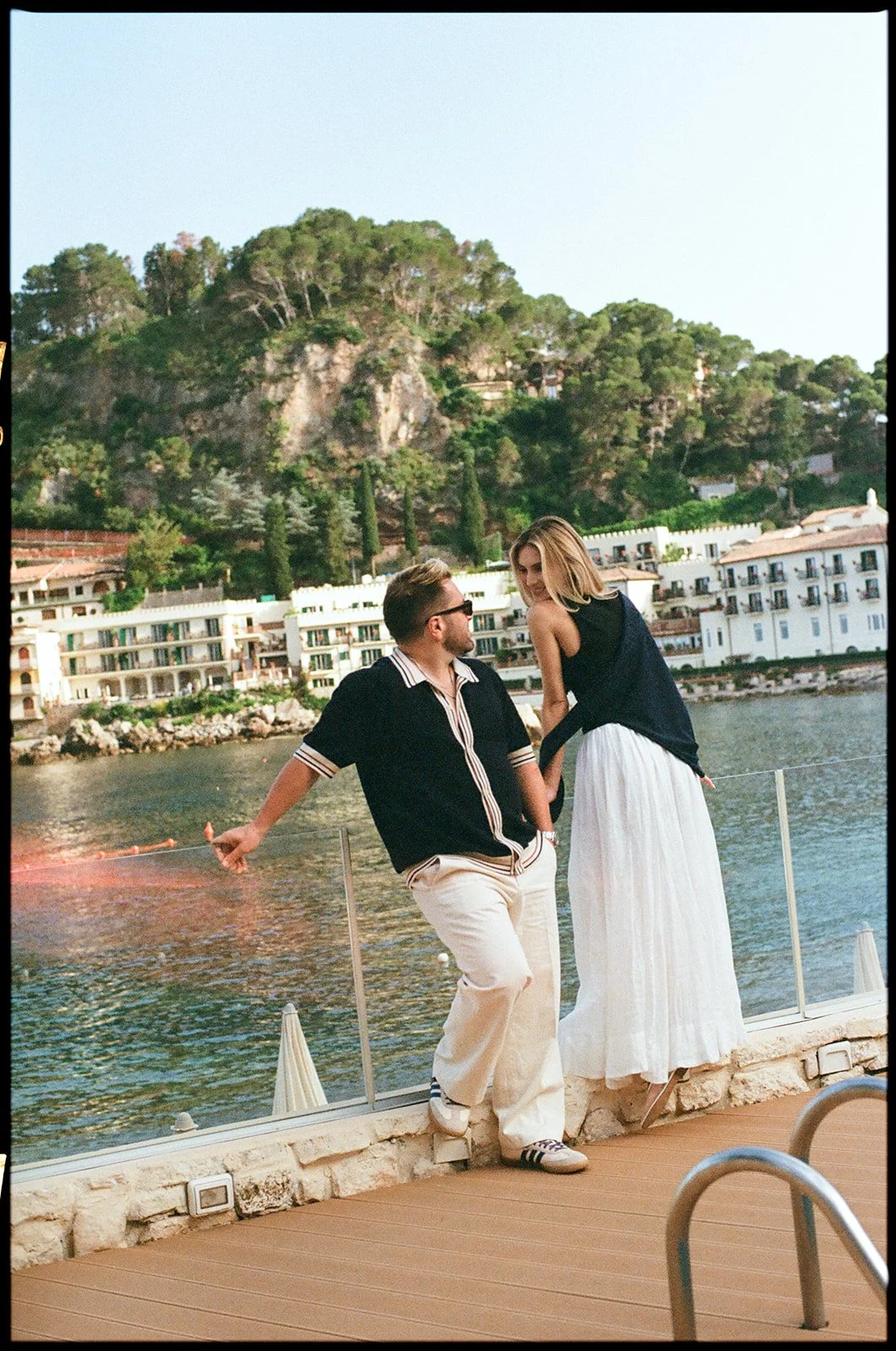 A man and woman standing by the water's edge on a dock, facing each other and holding hands with a scenic hillside and white buildings in the background.