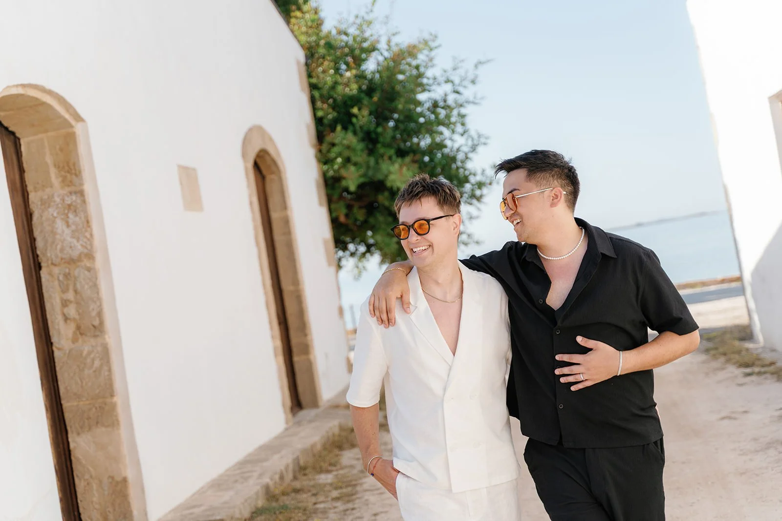 Two men walking arm-in-arm on a sunny beachside path, smiling and wearing sunglasses, with white buildings and the sea in the background.
