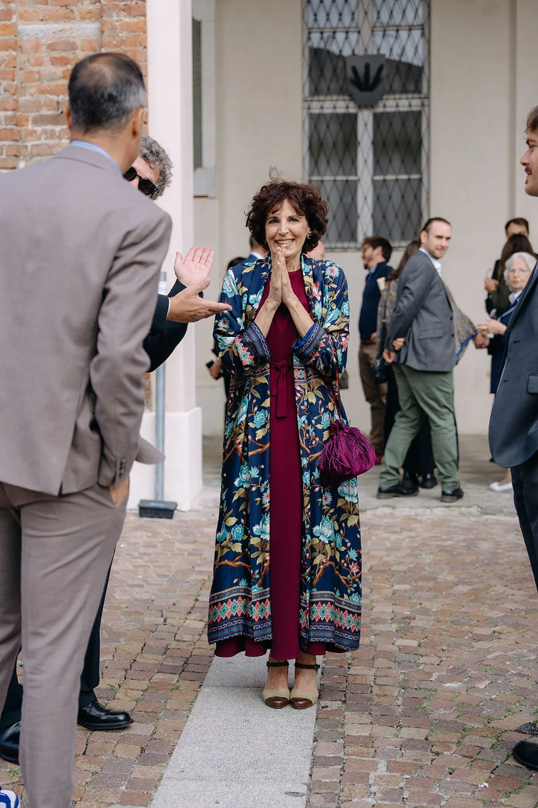 A woman in a long, colorful floral coat and burgundy dress stands on a cobblestone street, smiling with her hands together in a gesture of gratitude, while people in suits converse around her.