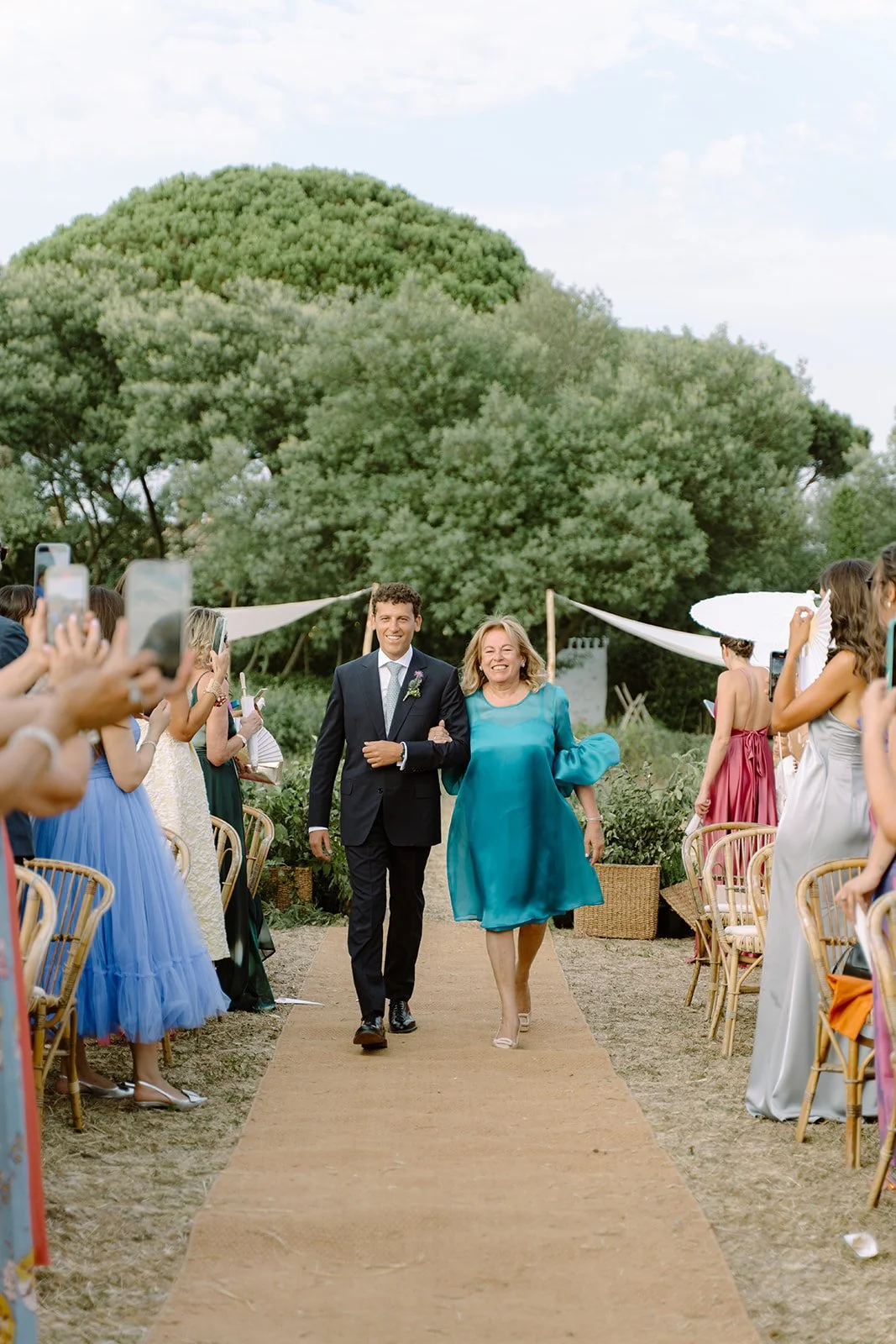 A woman in a teal dress walking down an outdoor aisle with a man in a suit, surrounded by people taking photos at a wedding ceremony outdoors with trees in the background.