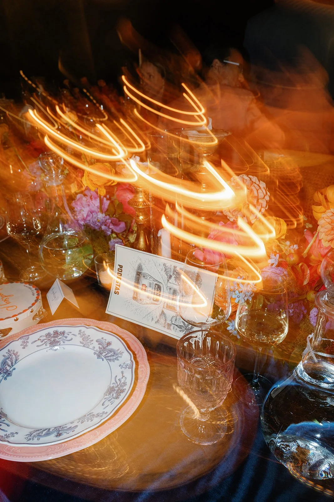 A table set for a celebration with a floral centerpiece, plates, glasses, and a menu card. The long exposure photo creates swirling light trails above the table.