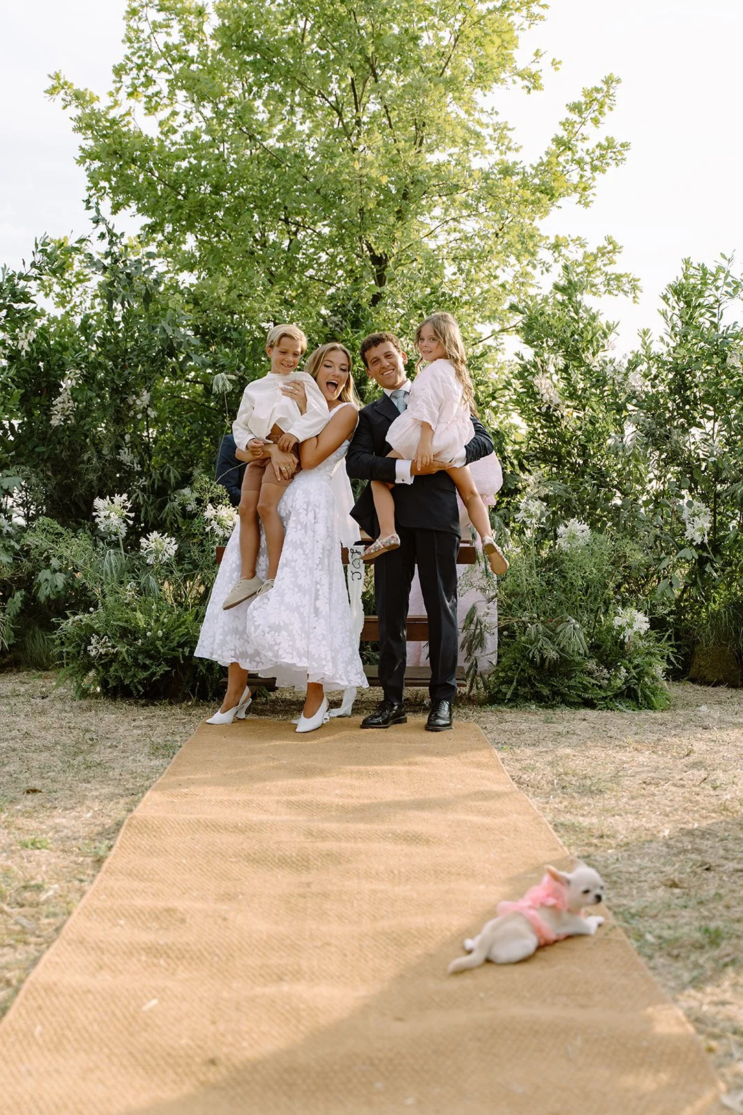 A couple dressed in wedding attire, the bride in a white lace dress and the groom in a black suit, standing on a beige carpet outside with three children. The children, two girls and a boy, are sitting on the couple's legs. The background features gr