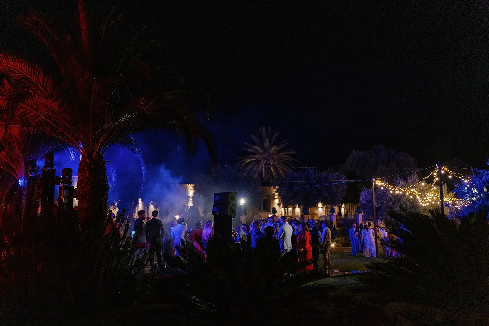 Nighttime outdoor celebration with a crowd of people, string lights, fireworks, and palm trees.