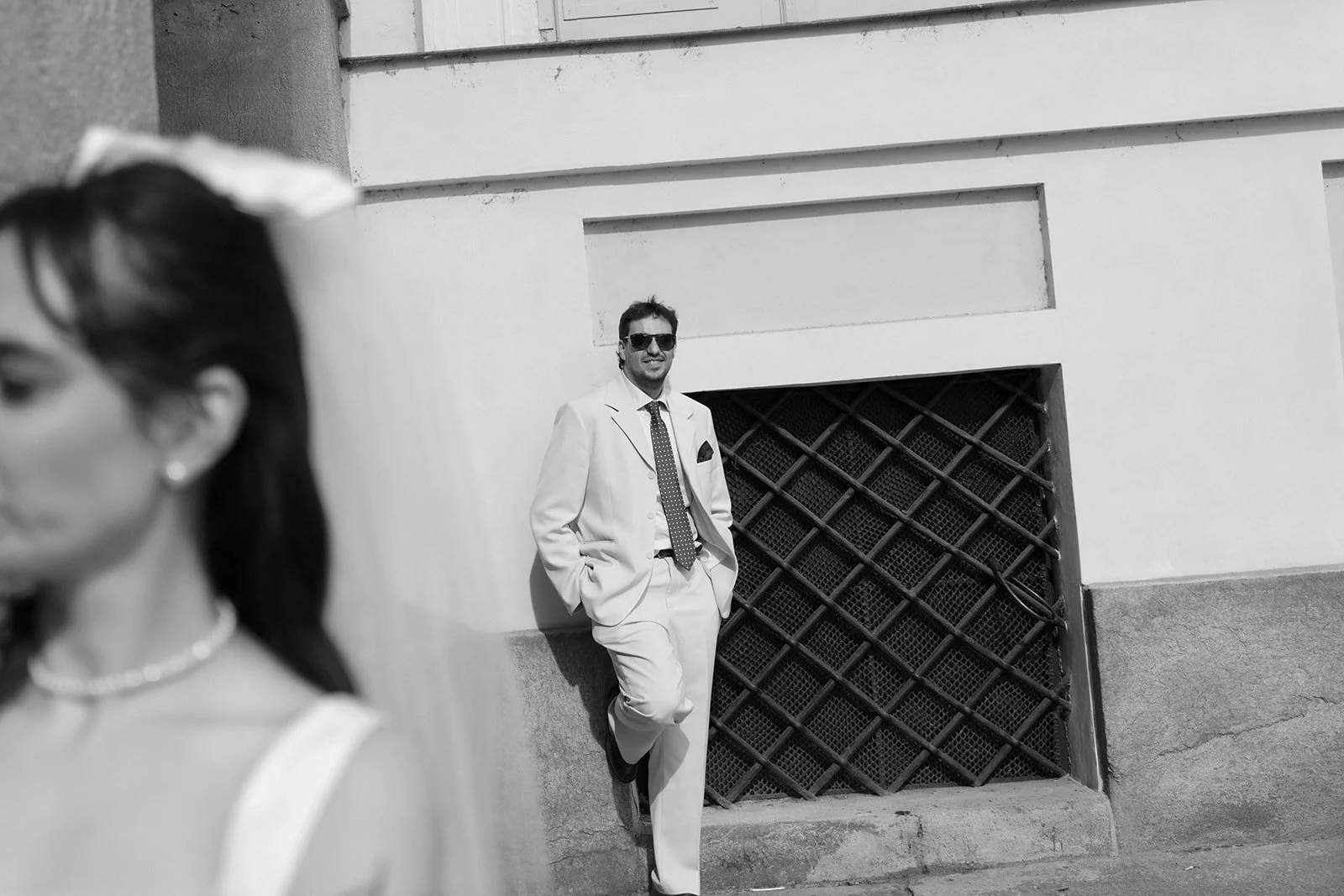 A man in a white suit with a tie and sunglasses leaning against a wall outside, with a woman with dark hair and pearl necklace in the foreground.