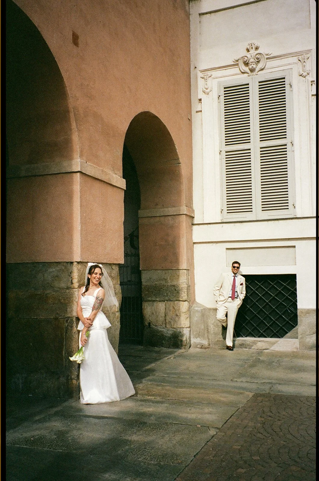 A bride in a white wedding dress smiling and holding a bouquet of flowers, standing near an archway in an old building, with a groom in a light-colored suit and sunglasses leaning against the wall nearby.