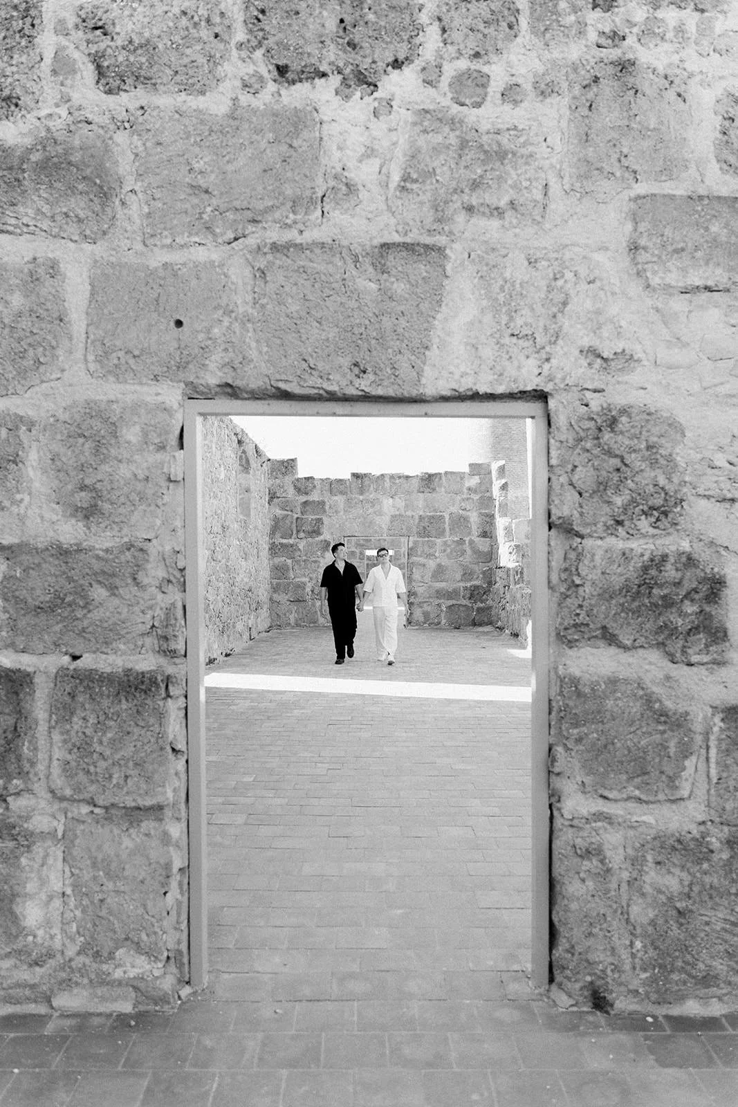 Two people walking hand in hand through a stone structure, with one person dressed in black and the other in white, viewed through a rectangular opening in a stone wall.