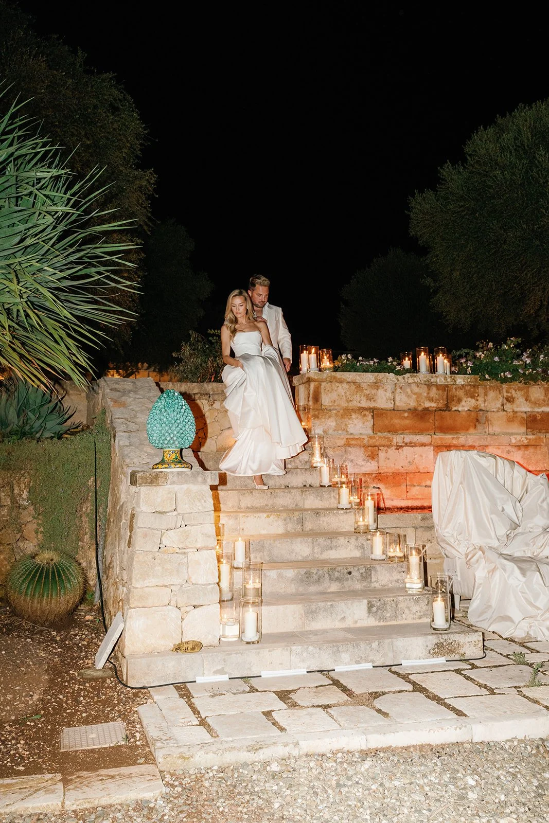 A bride in a white wedding gown and a groom in a white suit sitting on outdoor stone stairs at night, surrounded by candles in glass holders, with trees and a brick wall in the background.